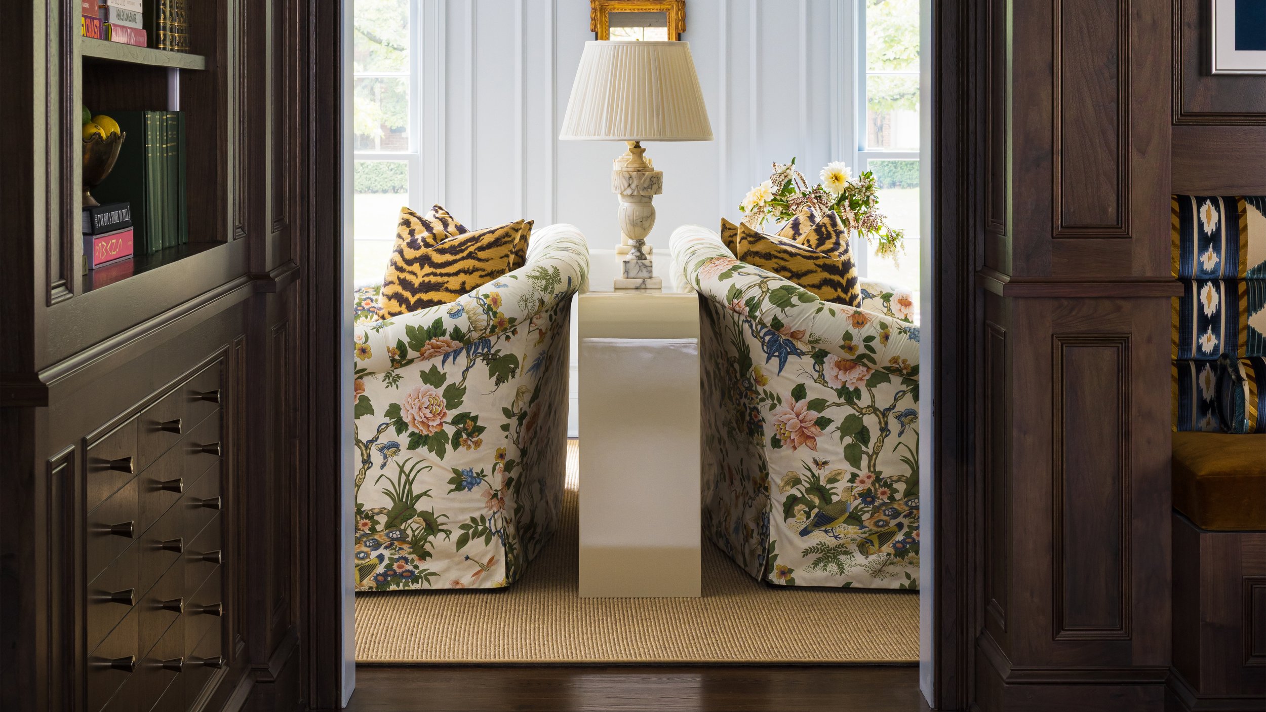 View from a doorway into a sunlit sitting area with floral and animal print armchairs, a marble table with a large lamp, and a window with white curtains.
