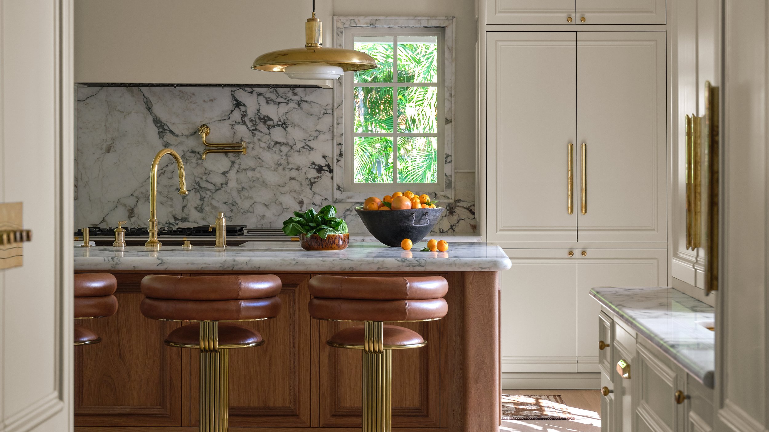 A kitchen with white marble countertops and backsplash, gold fixtures, a window with greenery outside, and a central island with brown stools. There is a bowl of oranges and some greenery on the island.