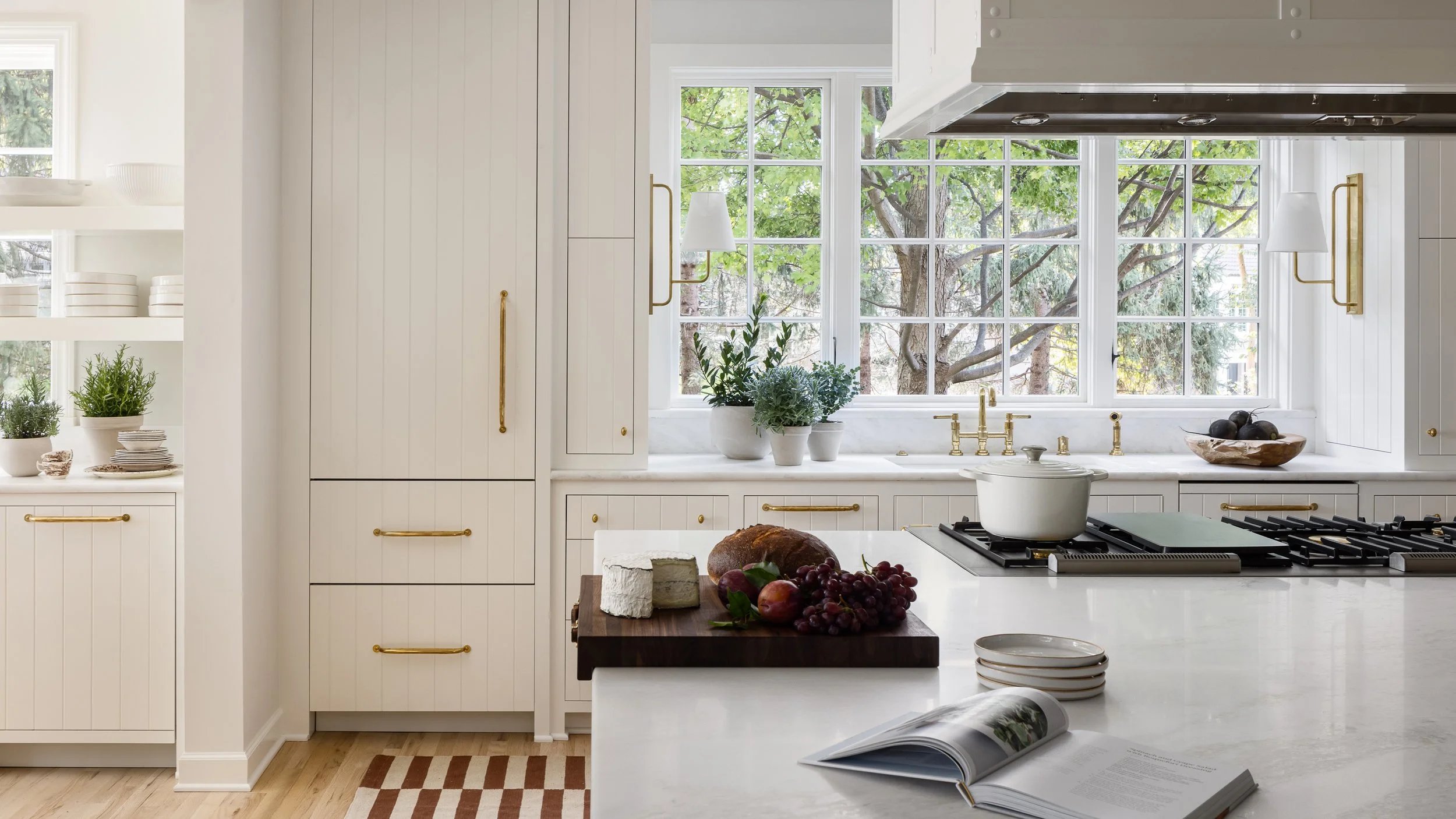 A bright white kitchen with large windows overlooking trees. The kitchen has white cabinets with gold handles, a marble countertop, potted plants, a bowl of black onions, and a cookbook open on the counter.
