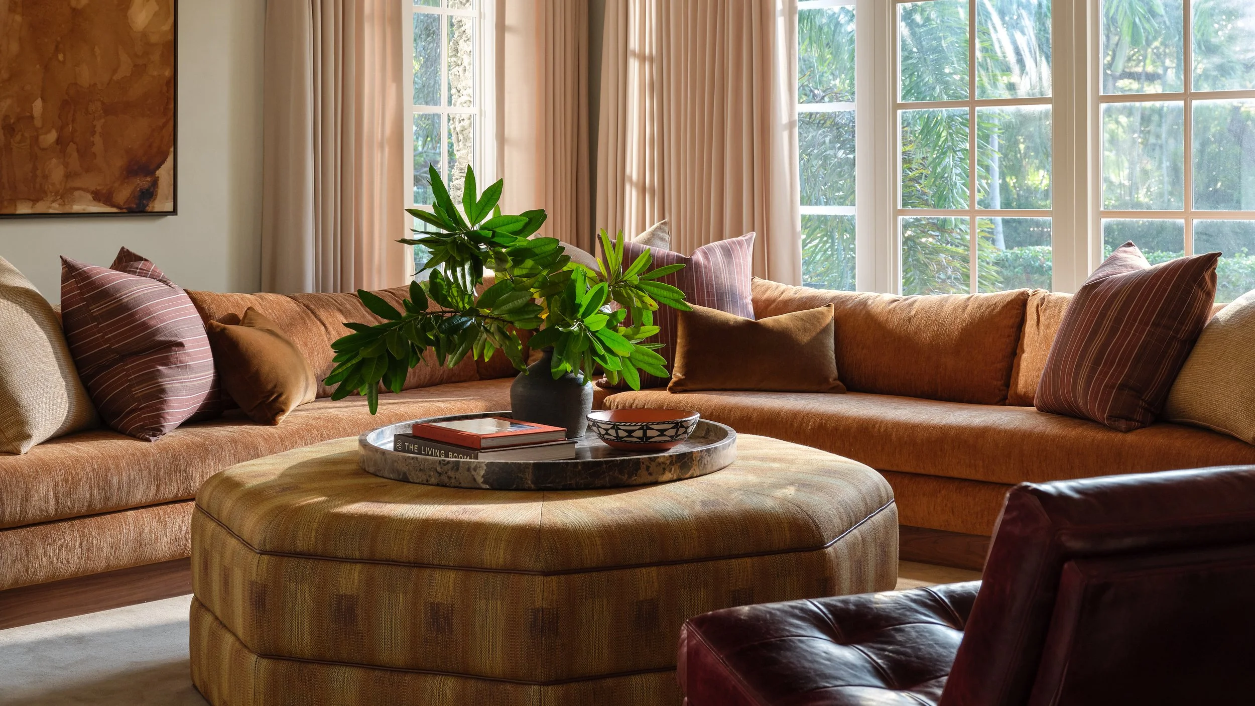 A cozy living room with large windows, curtains, a sectional sofa with multiple cushions, a round ottoman, a marble tray with books and a bowl, and a large green plant in a vase.
