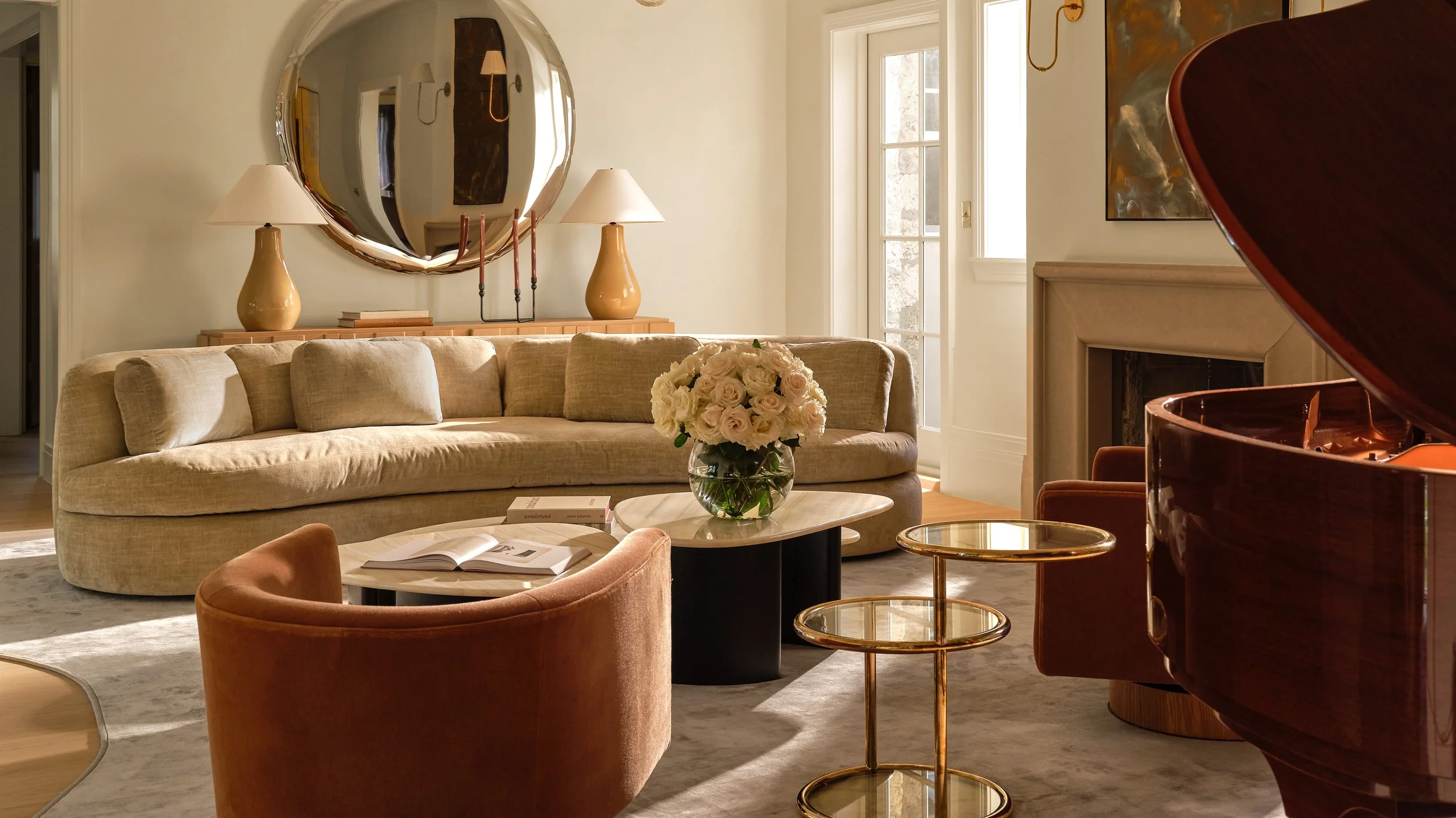 A living room with a beige sofa, a round coffee table with a flower arrangement, and a grand piano. There are lamps, books, a mirror, and art on the walls, with large windows allowing natural light.