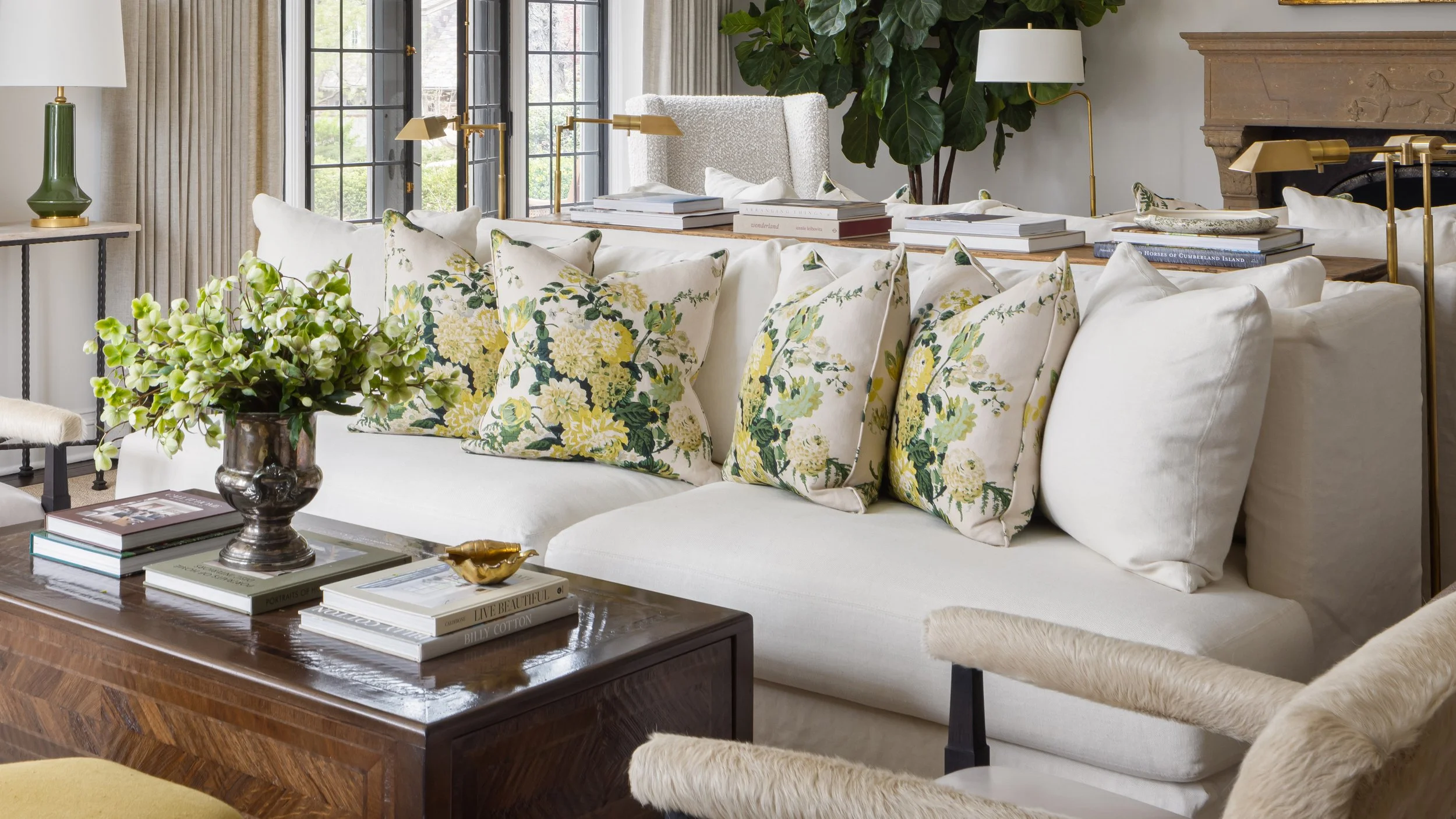 Living room with white sofa decorated with pillows featuring yellow and green floral patterns, a wooden coffee table with books and a flower arrangement, black window frames, beige curtains, and a fireplace with green plants and lamps nearby.
