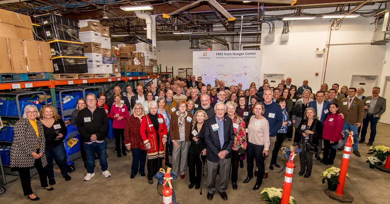 Group of people gathered inside a warehouse with shelves filled with boxes and containers, in front of a whiteboard that reads 'FREE from Hunger Center'.