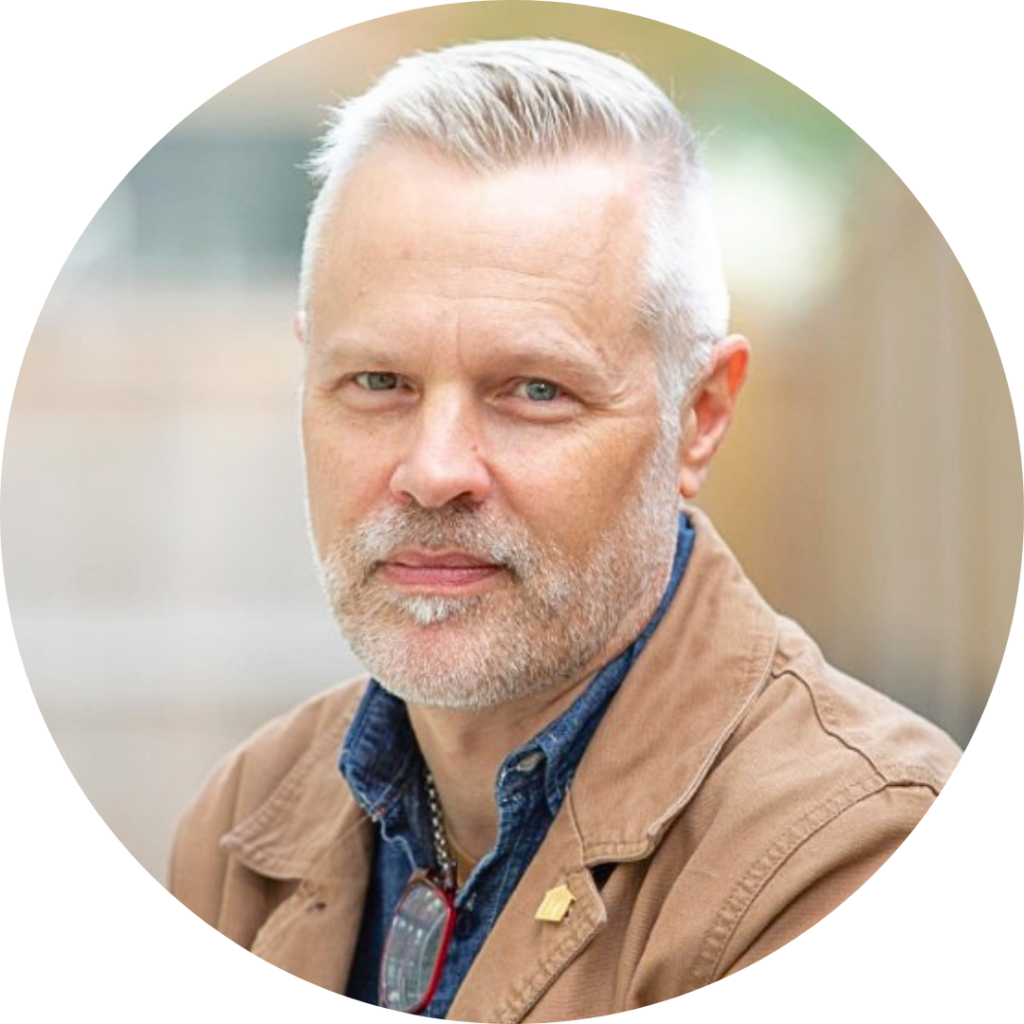 Close-up portrait of a middle-aged man with light-colored hair and beard, wearing a brown jacket and blue shirt, outdoors during daytime.