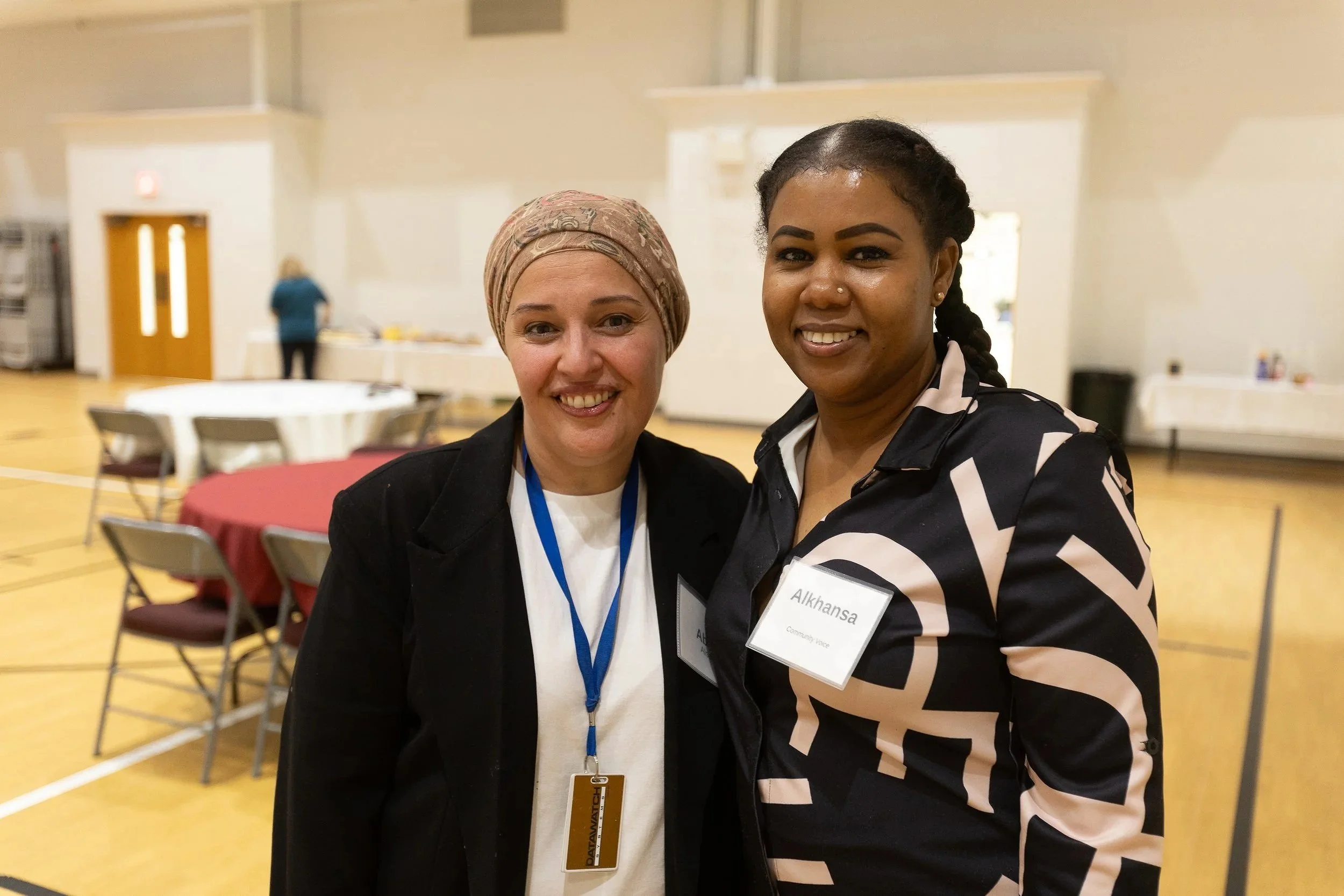 Two women standing close together in a large indoor space, smiling at the camera. One woman has light skin, is wearing a beige headscarf, a black jacket, and a white shirt. The other woman has darker skin, braided hair, and is wearing a black and pink patterned blazer. Both women appear to be at a professional or community event, as indicated by their name tags.