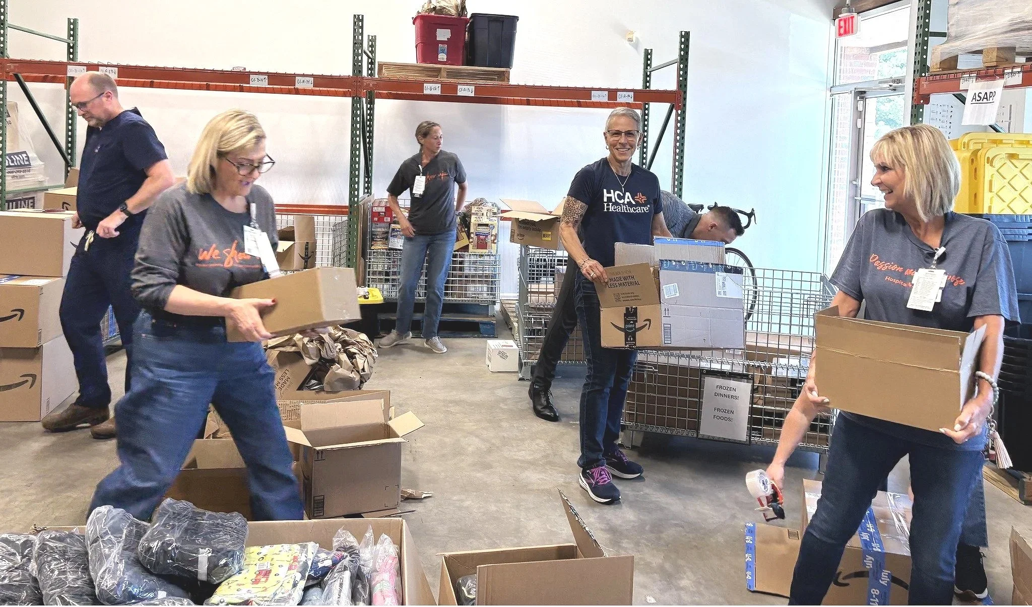 Group of people working together to pack and organize boxes in a warehouse or donation center. They are handling cardboard boxes with supplies, and some are smiling and engaging in tasks.
