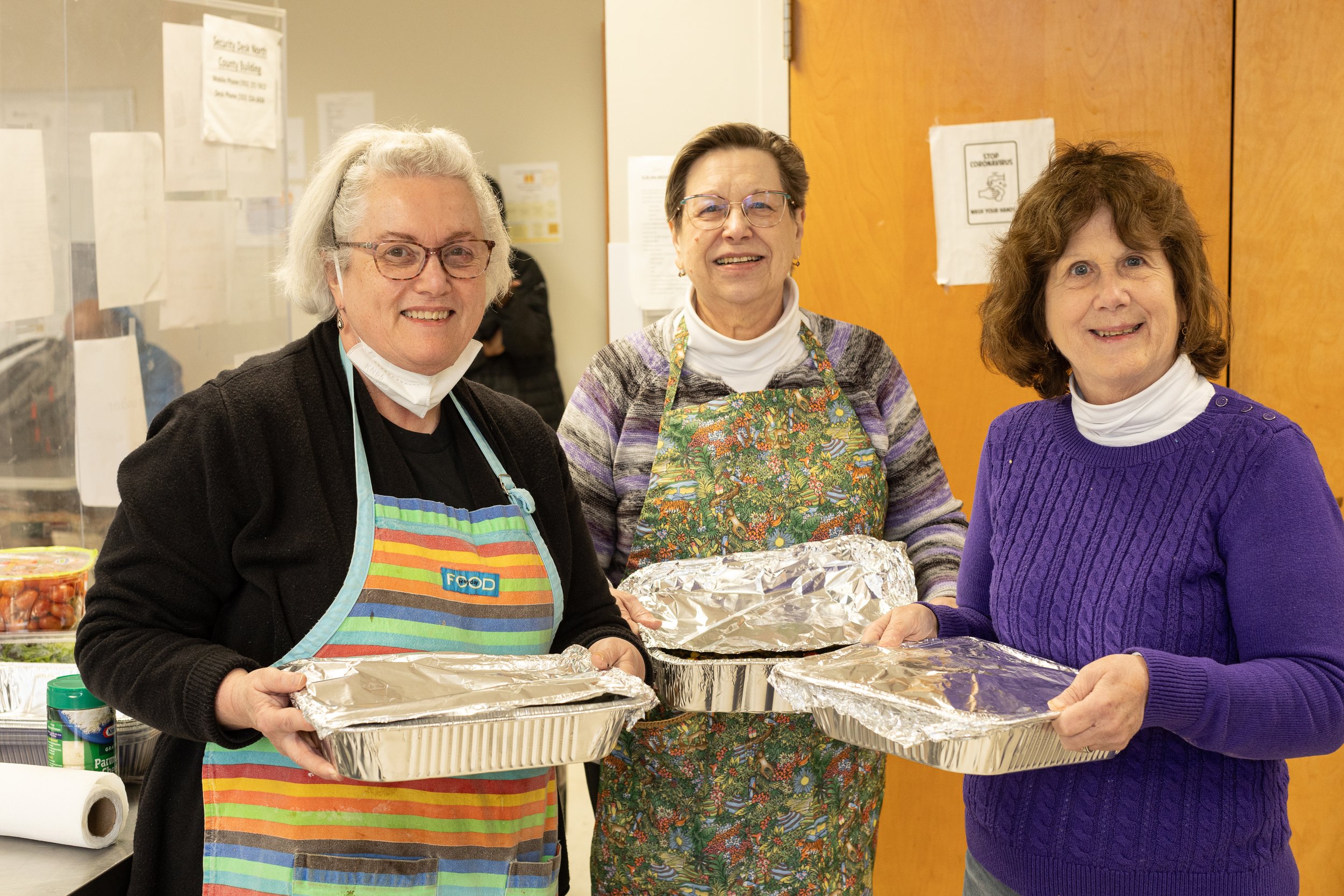 Three women standing behind a table with aluminum foil trays, smiling at the camera. They are in a kitchen or community room, wearing aprons and casual clothing.