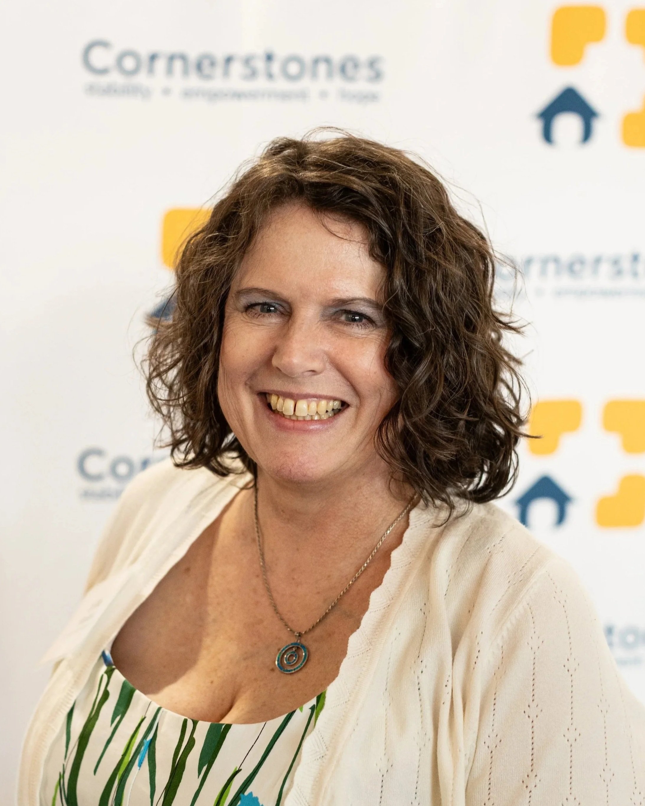 Smiling woman with curly brown hair wearing a cream cardigan and a necklace with a circular pendant, standing in front of a backdrop with the words "Cornerstones" and icons of a house and puzzle pieces.