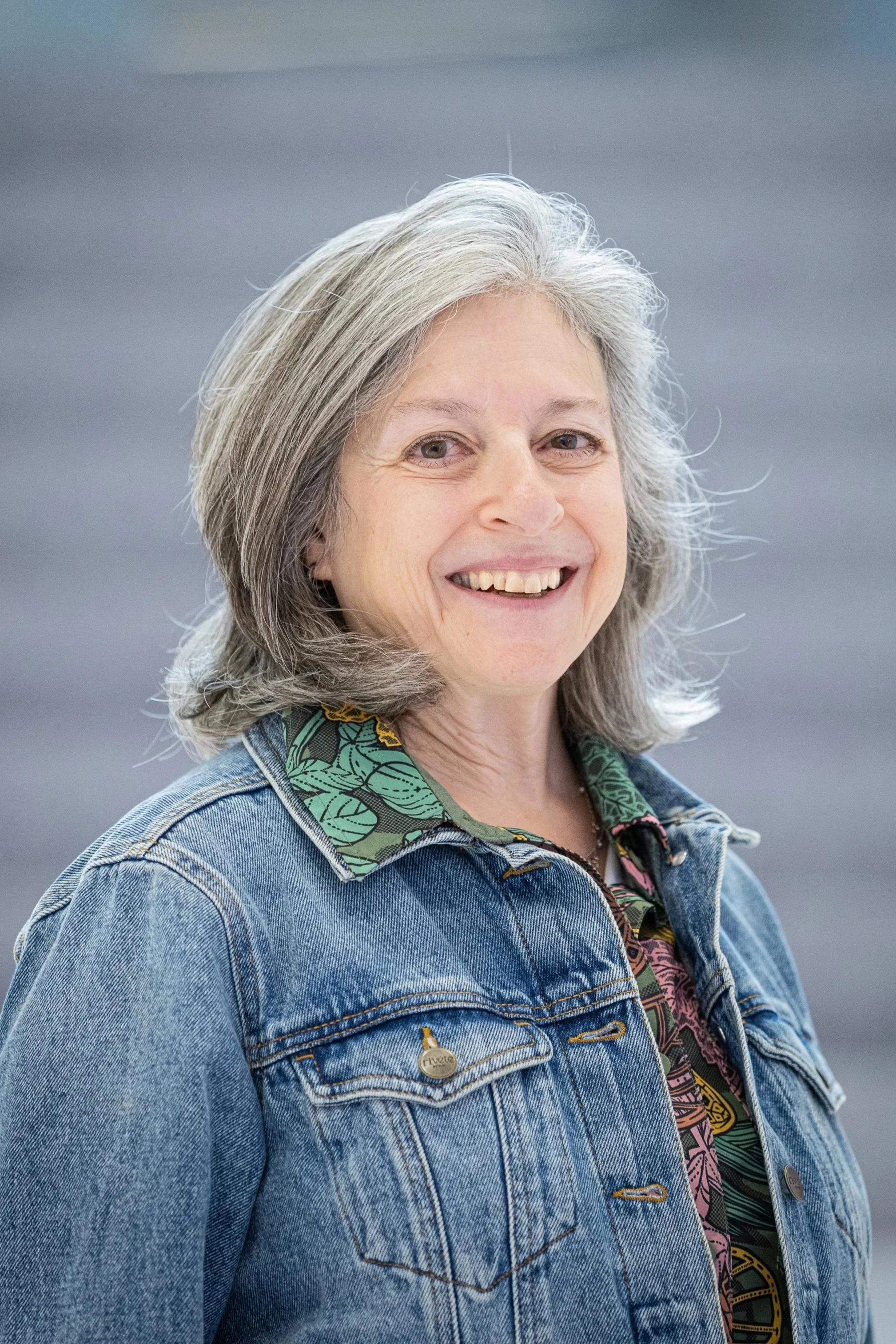 Smiling woman with gray hair wearing a denim jacket and patterned shirt, standing outdoors against a blurred gray background.