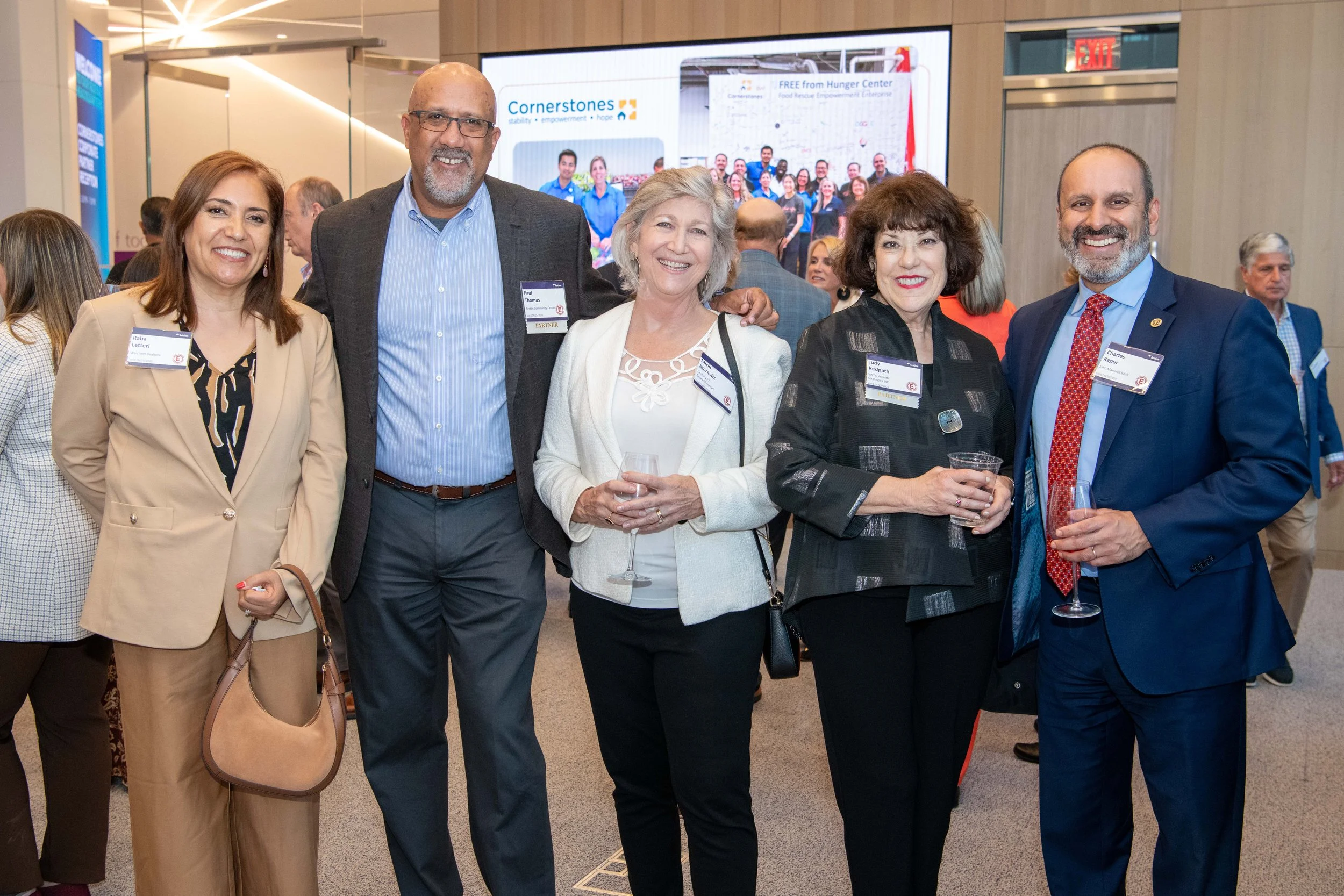 Group of five professionally dressed people, three women and two men, smiling at a networking event or conference, with a large screen in the background displaying the logo 'Cornerstones' and a group photo.