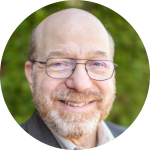 A smiling man with glasses and a beard, wearing a light-colored shirt.
