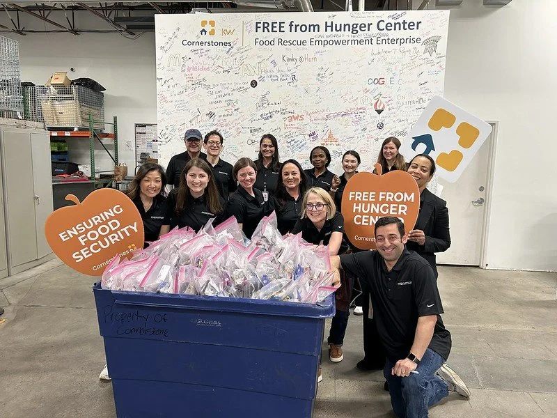 Group of employees standing behind a large blue recycling bin filled with plastic bags. They are inside a facility with shelves and equipment, holding signs that say "Ensuring Food Security" and "Free from Hunger Center." A whiteboard on the wall behind them has writing and signatures, and a large sign above reads "Free from Hunger Center," indicating the group's involvement in a food rescue and empowerment initiative.