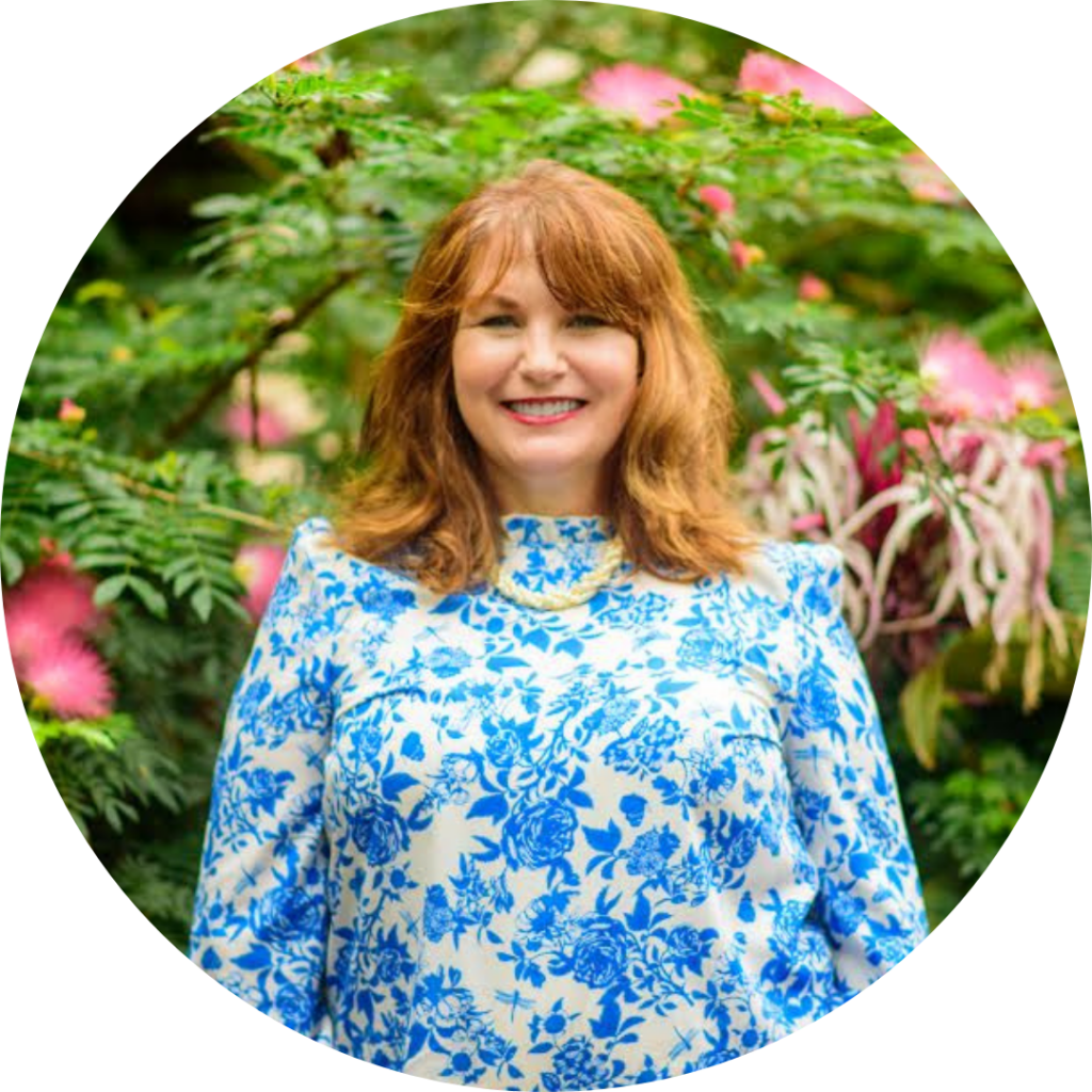 A woman with red hair smiling in front of green foliage and pink flowers.