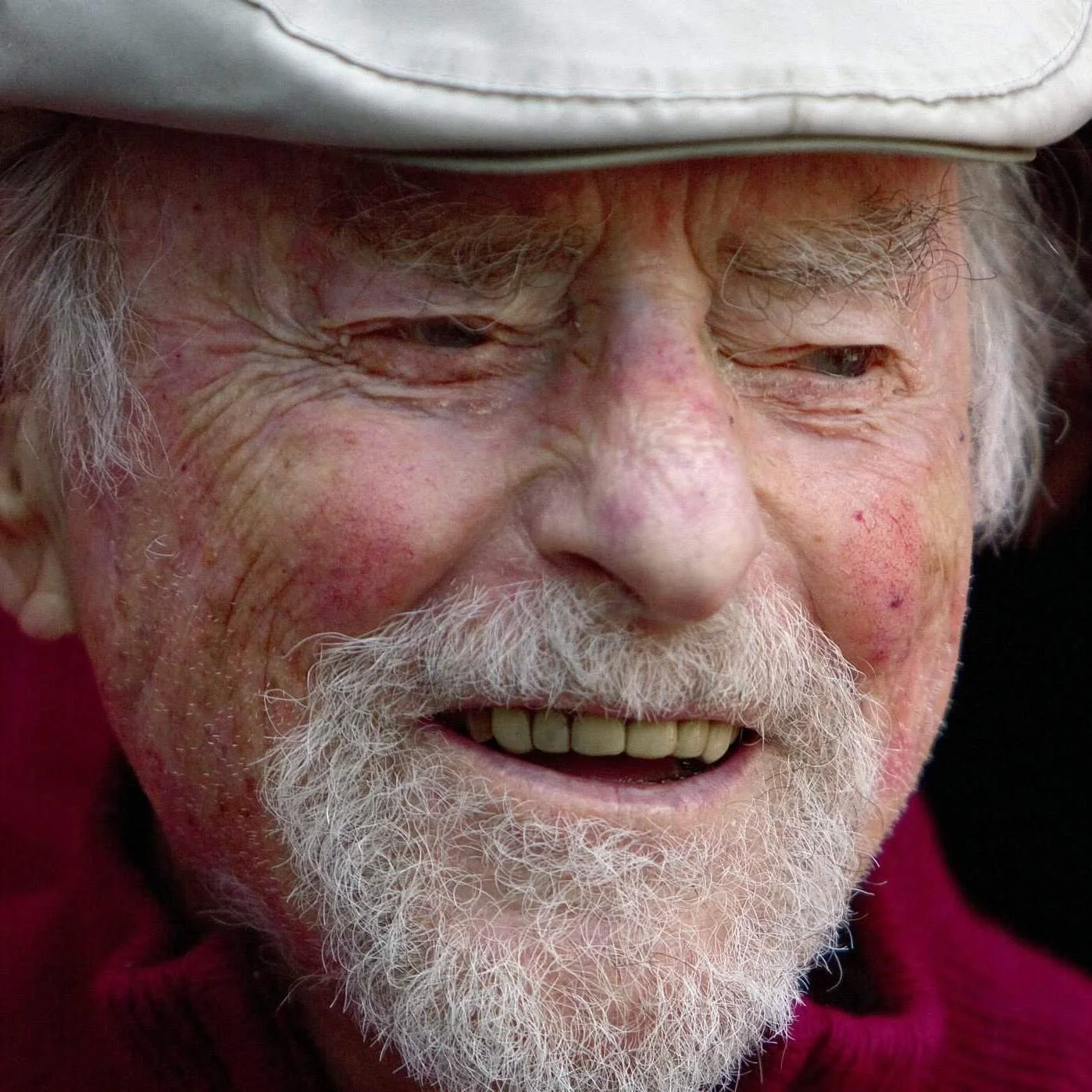 A close-up of the founder of Reston, Robert E. Simon, as an elderly man with white hair, a white beard, and mustache, wearing a light-colored hat and a red shirt, smiling with his eyes partially closed.