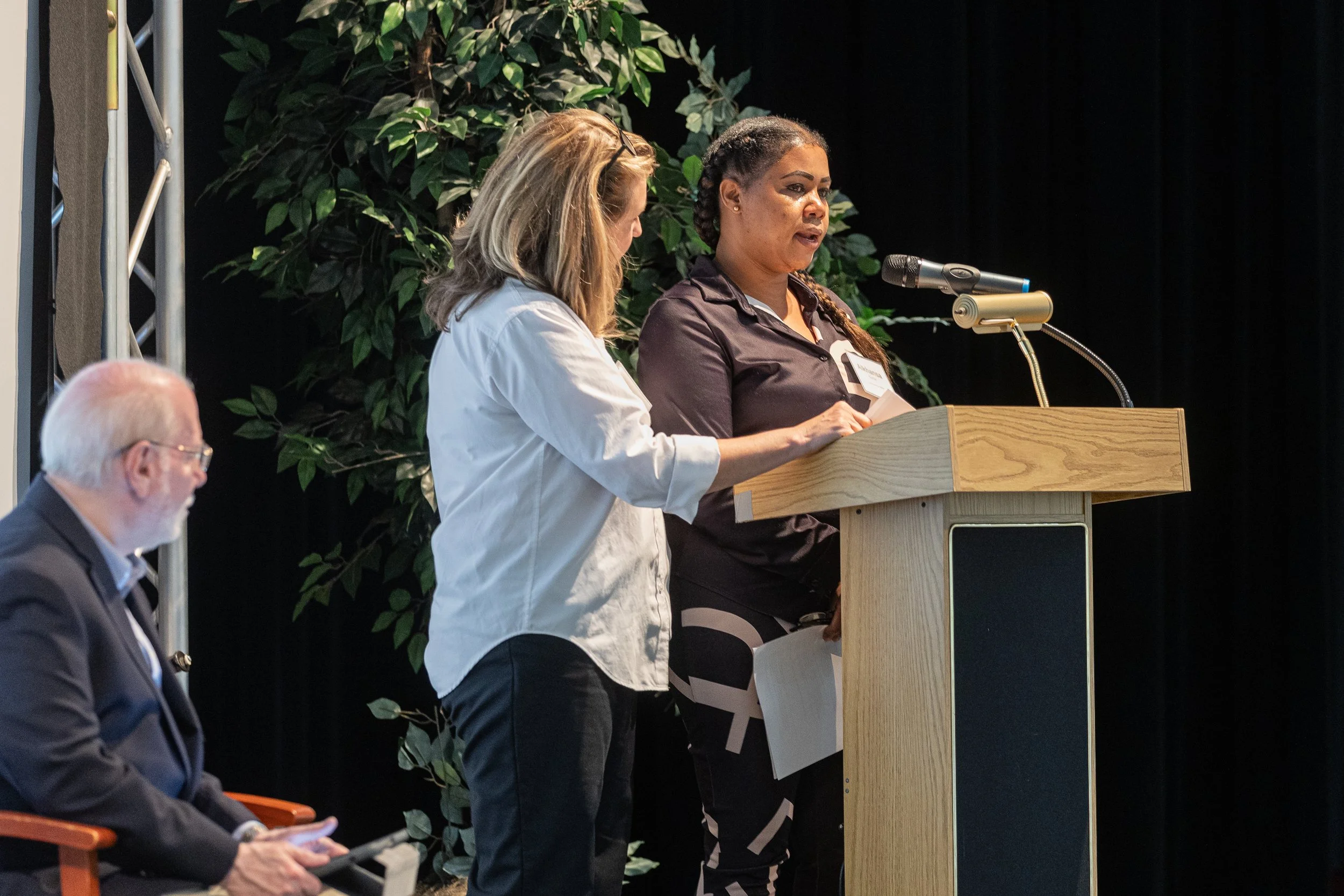 Two women stand at a podium, one speaking into a microphone while the other looks on. A man sits in the foreground on the left side, wearing glasses and a suit. There are green leafy plants behind the women and a black curtain backdrop.