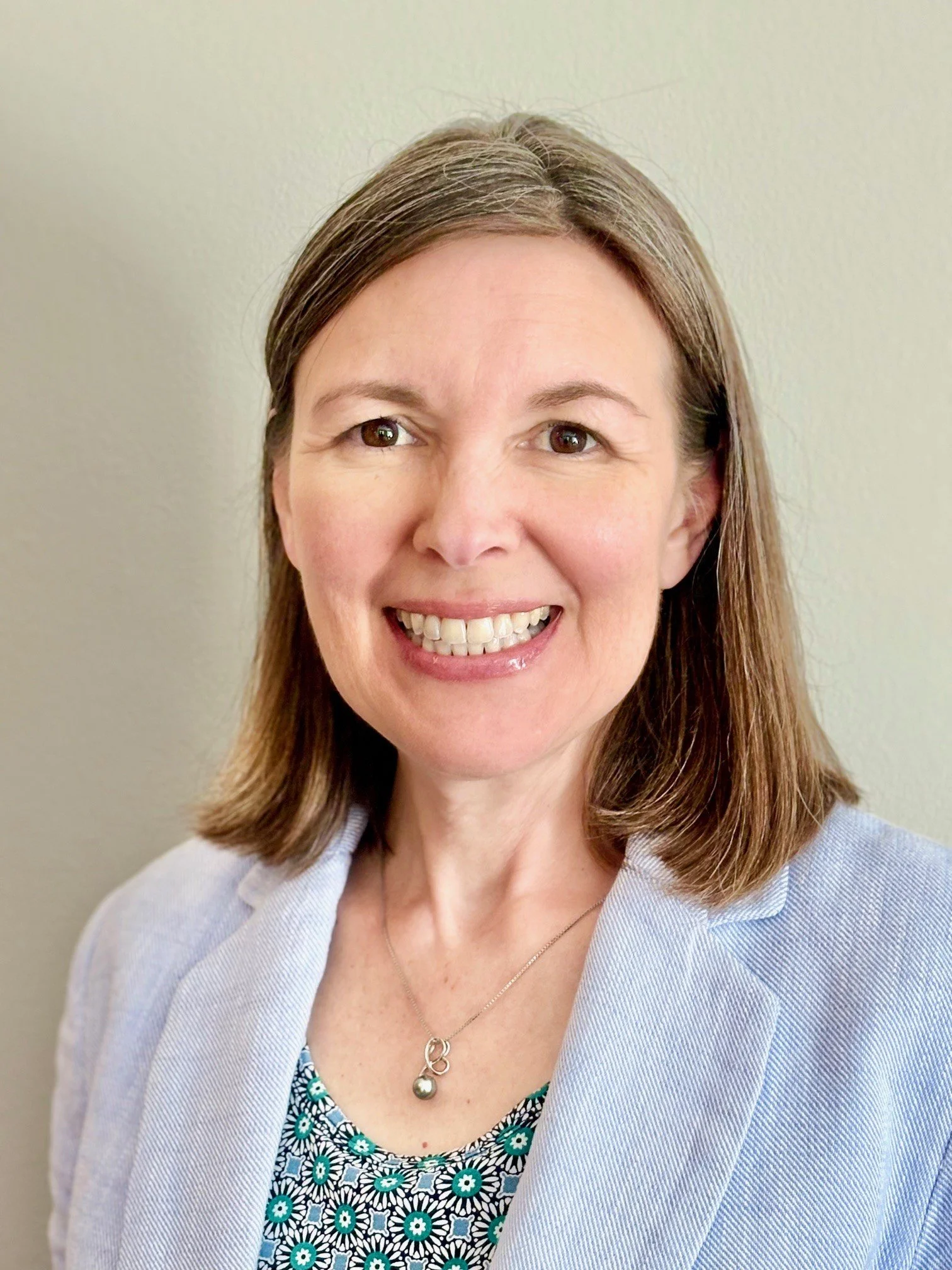 A woman with shoulder-length brown hair, smiling, wearing a light blue blazer over a patterned top, and a necklace with a pendant, standing against a plain beige background.