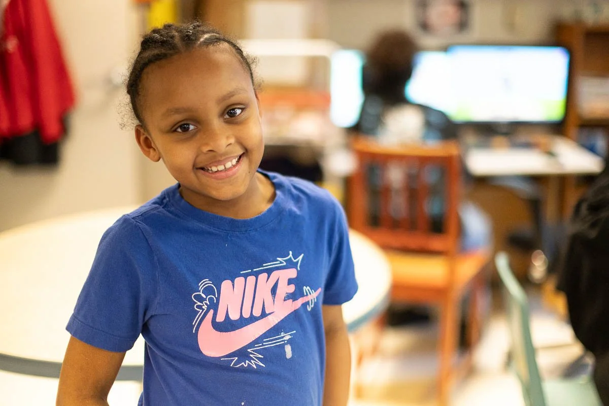 A smiling young girl with braids wearing a blue Nike t-shirt standing in a classroom or computer lab.