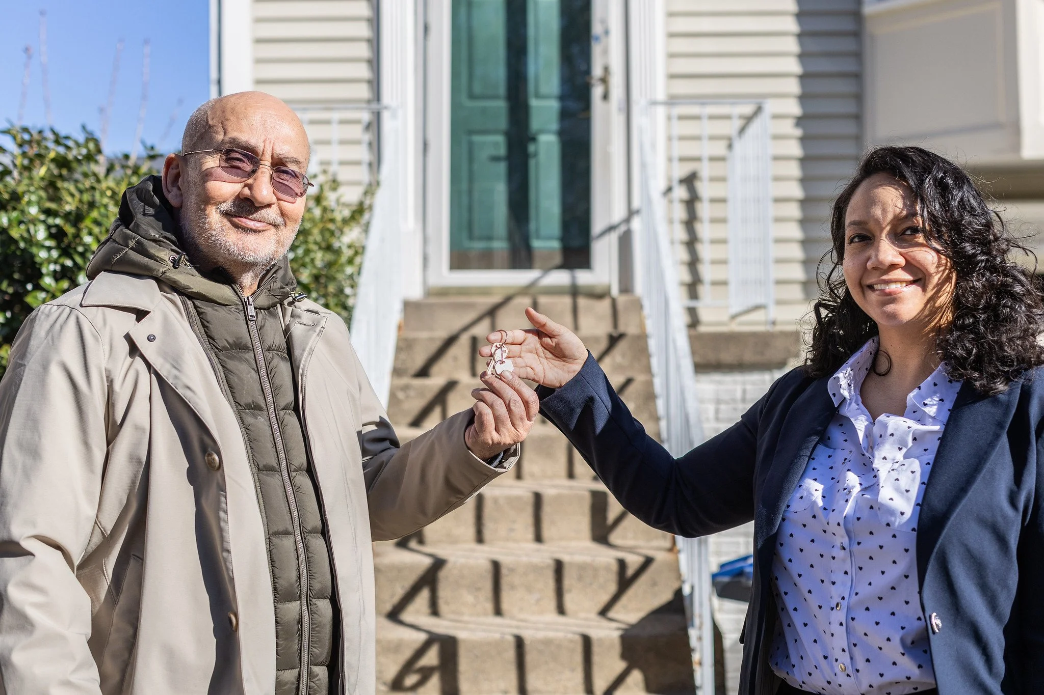 A man with glasses and a woman with curly hair are smiling and holding keys together in front of a house with stairs leading up to a door.