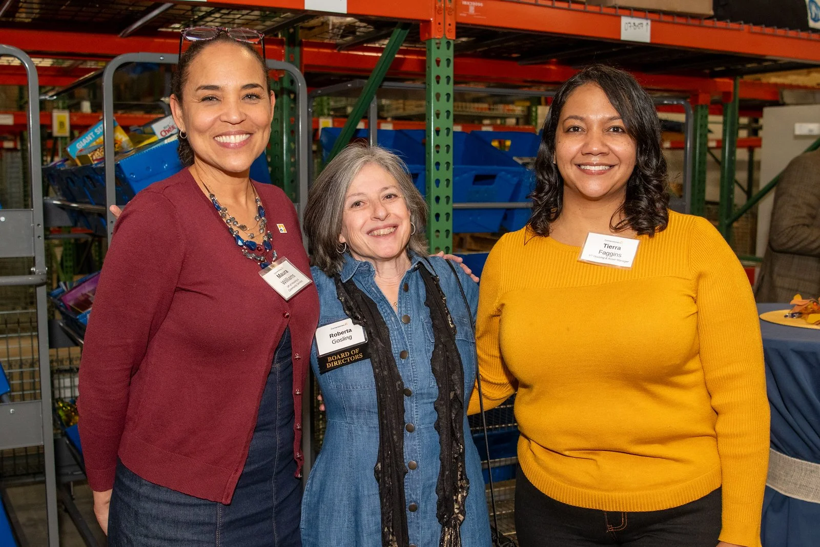 Three women standing together at a warehouse event, smiling at the camera. They are wearing name tags and casual clothing, with shelves and storage bins in the background.