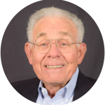 Portrait of an elderly man with white hair and glasses, wearing a dark suit and a light blue shirt, smiling against a dark gray background.