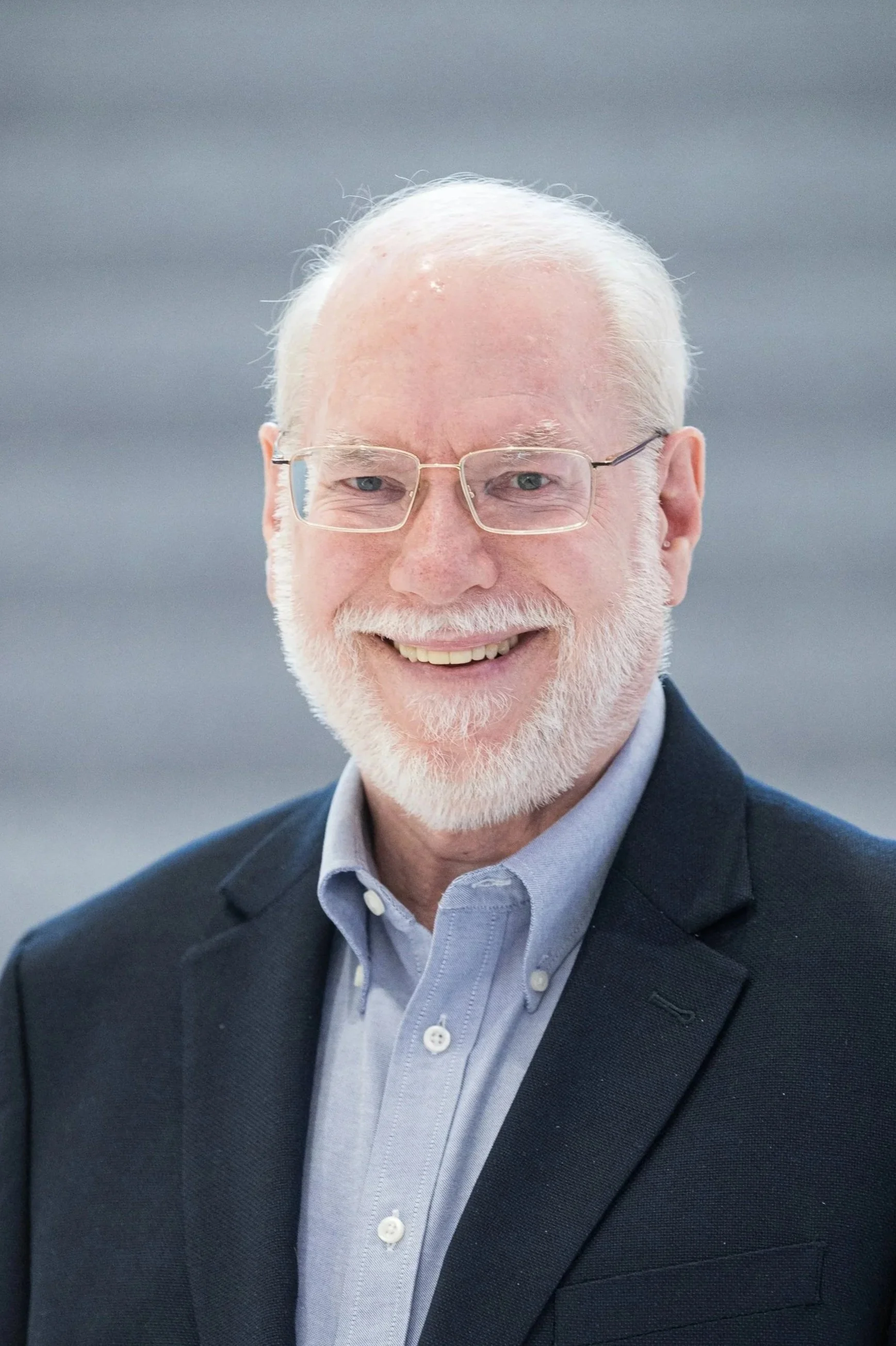 A smiling older man with white hair and a beard, wearing glasses, a light blue shirt, and a dark blazer, standing outdoors against a gray background.