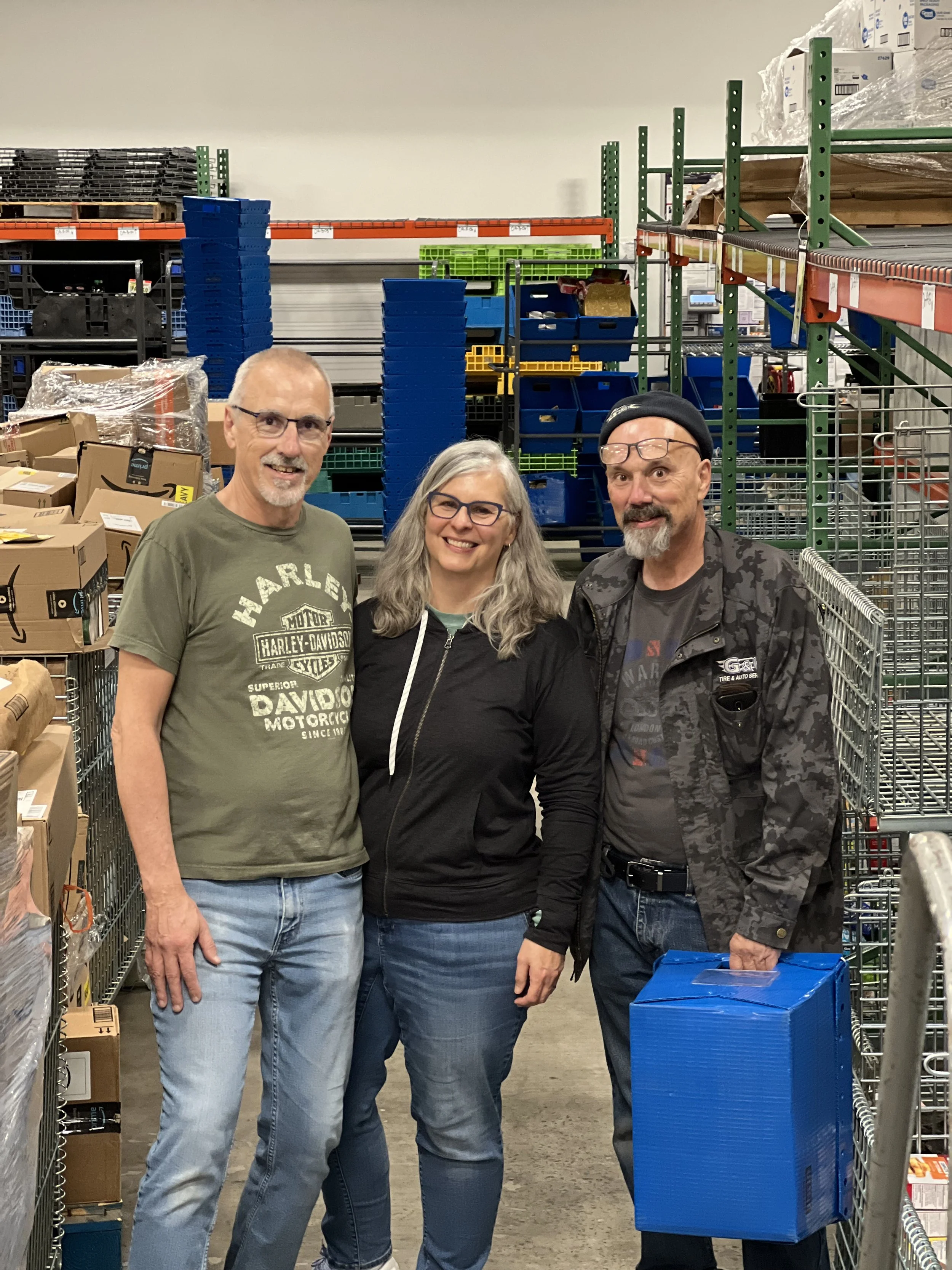 Three adult Cornerstones volunteers inside the Food Hub food rescue warehouse aisle, smiling at the camera, surrounded by shelves filled with boxes that they have been unpacking and sorting.