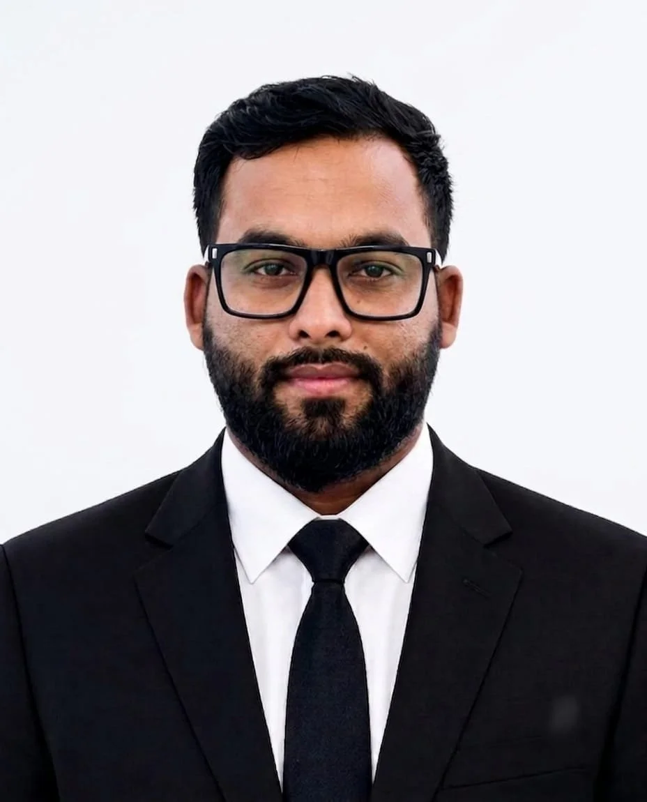 A man with black hair, beard, glasses, wearing a black suit, white shirt, black tie, against a plain white background.