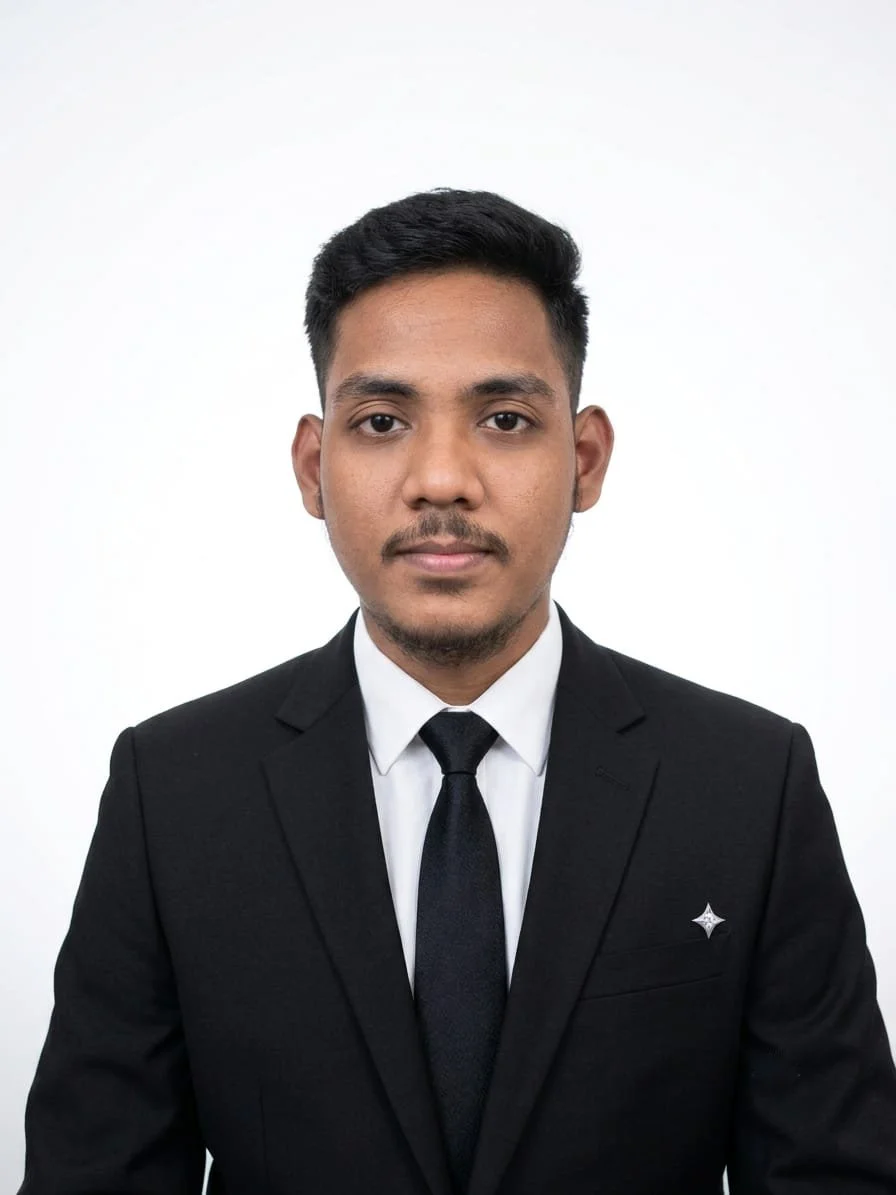 A young man with short black hair, wearing a black suit, white shirt, and black tie, posing against a plain white background.