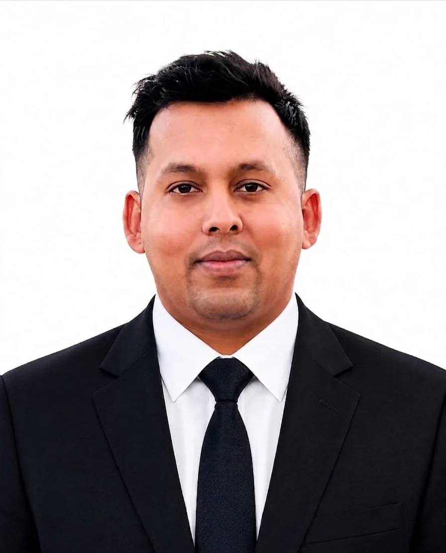 A man wearing a black suit, white shirt, and black tie facing the camera with a plain white background.