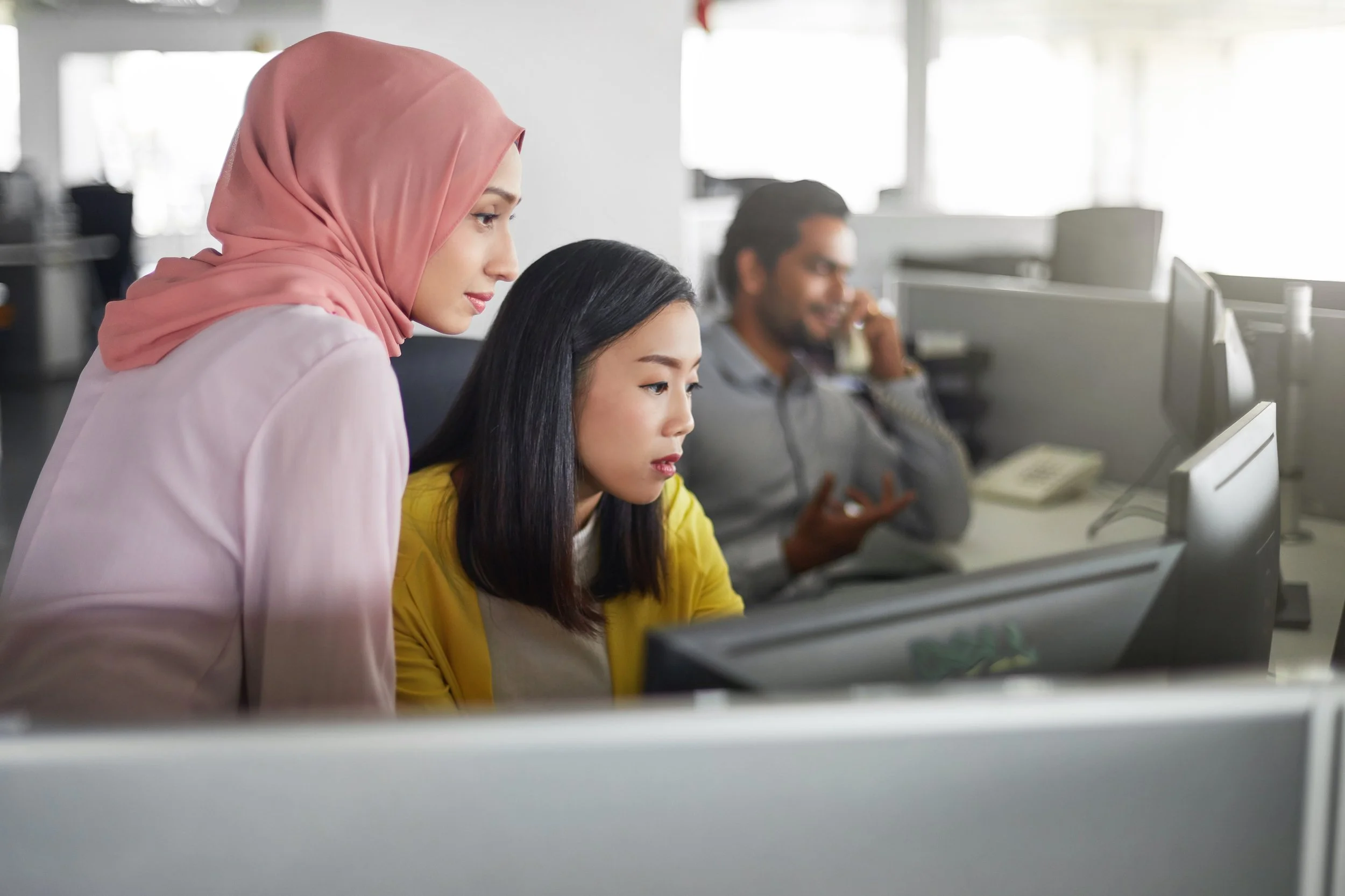 Three coworkers in an office, focused on their computers. One woman in a pink hijab is standing, looking at her monitor. Another woman in a yellow shirt is seated, also working on her computer. A man in a gray shirt is in the background, talking on the phone.