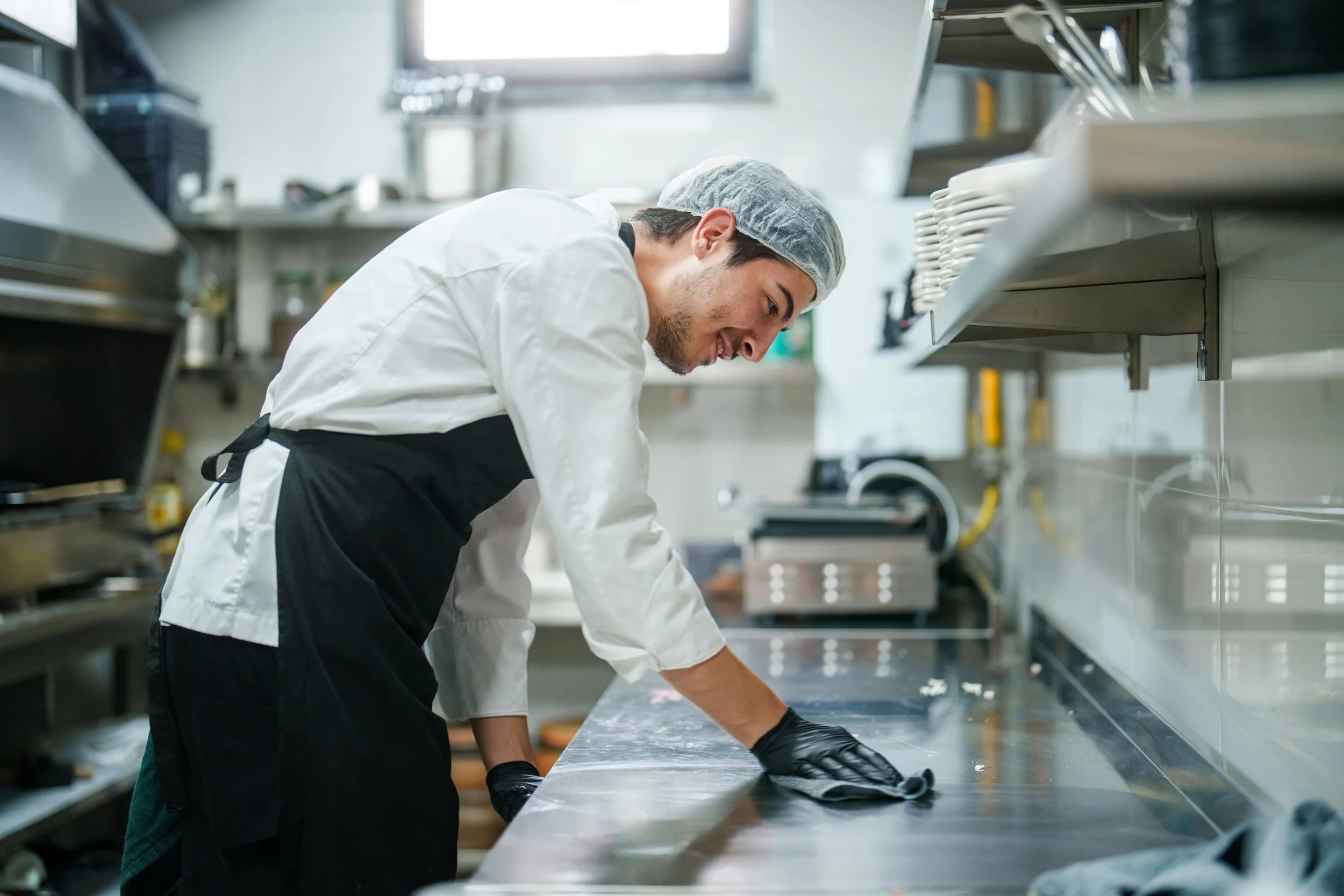 A male chef in a white uniform and a hairnet is cleaning a stainless steel countertop in a commercial kitchen.