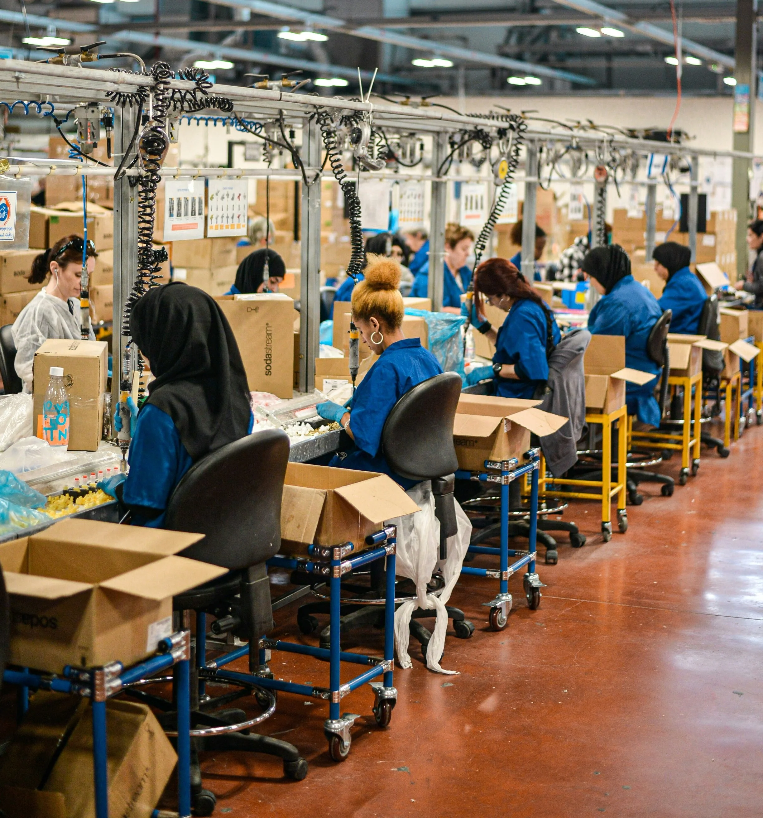Workers in a factory assembly line wearing blue uniforms and working with cardboard boxes. The factory has industrial lighting and metal workstations with coiled air hoses hanging above.