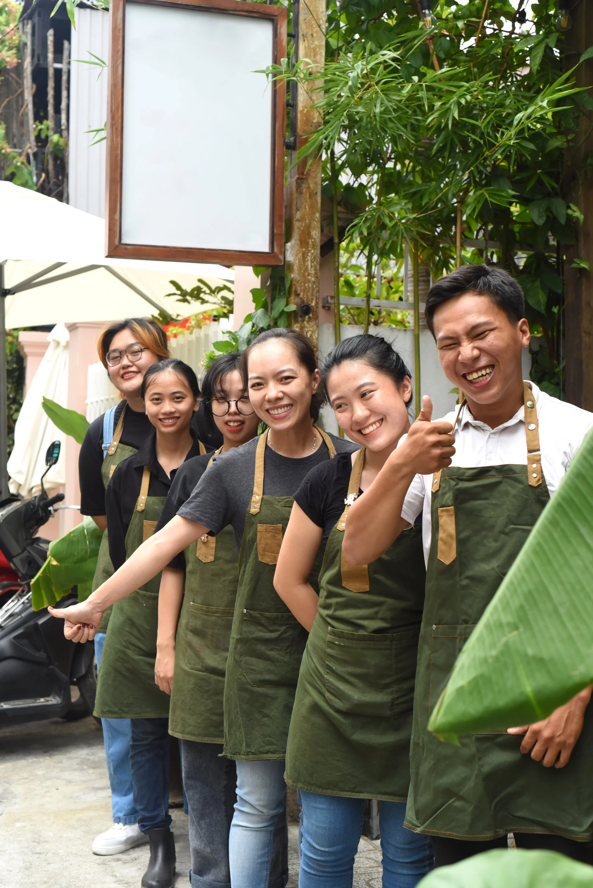 A group of six young adults standing in a line outdoors, wearing aprons and smiling. They are surrounded by green plants and appear to be at a gardening or outdoor event.