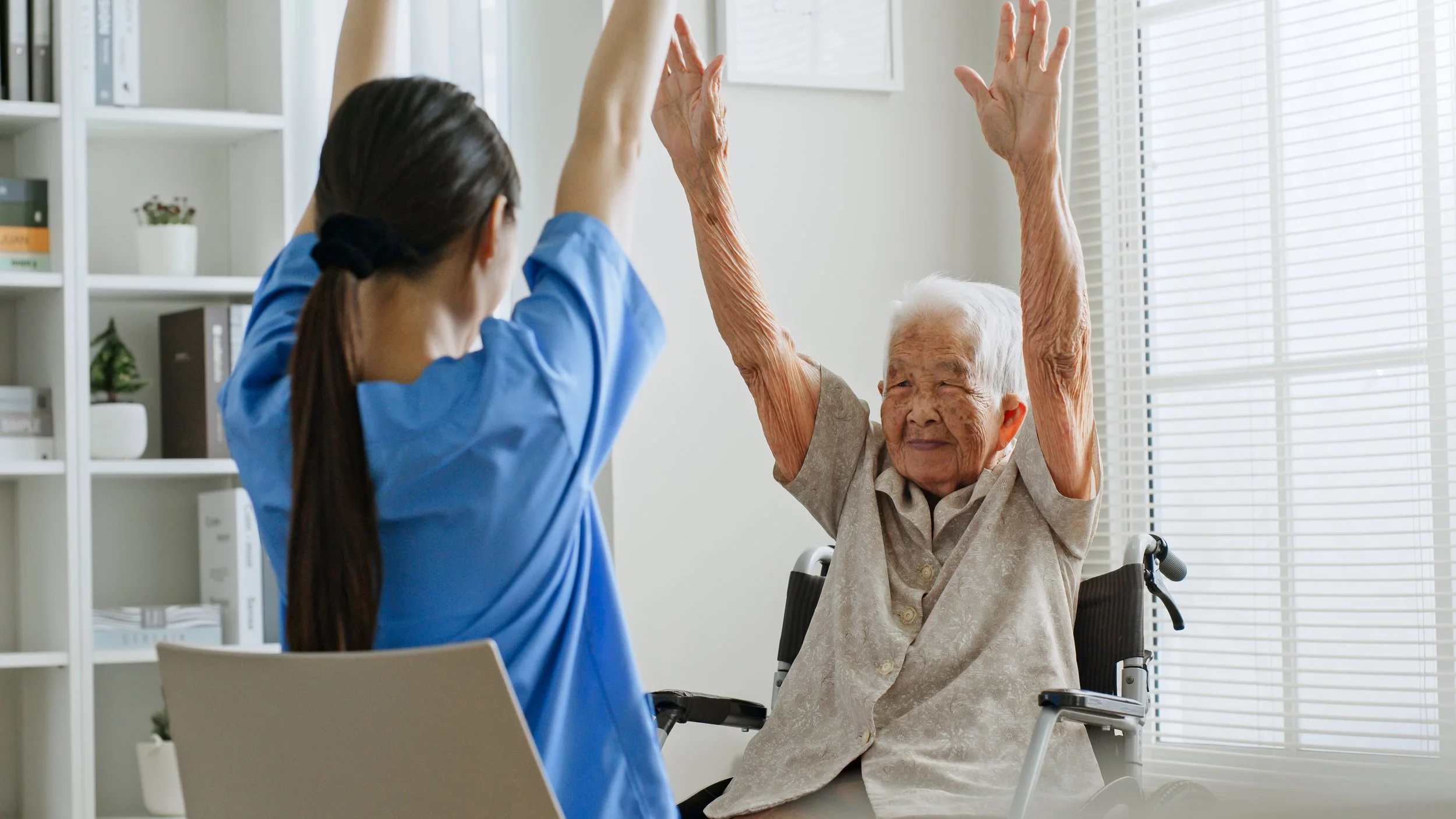 An elderly woman in a wheelchair doing exercise with a caregiver in a bright room with blinds.
