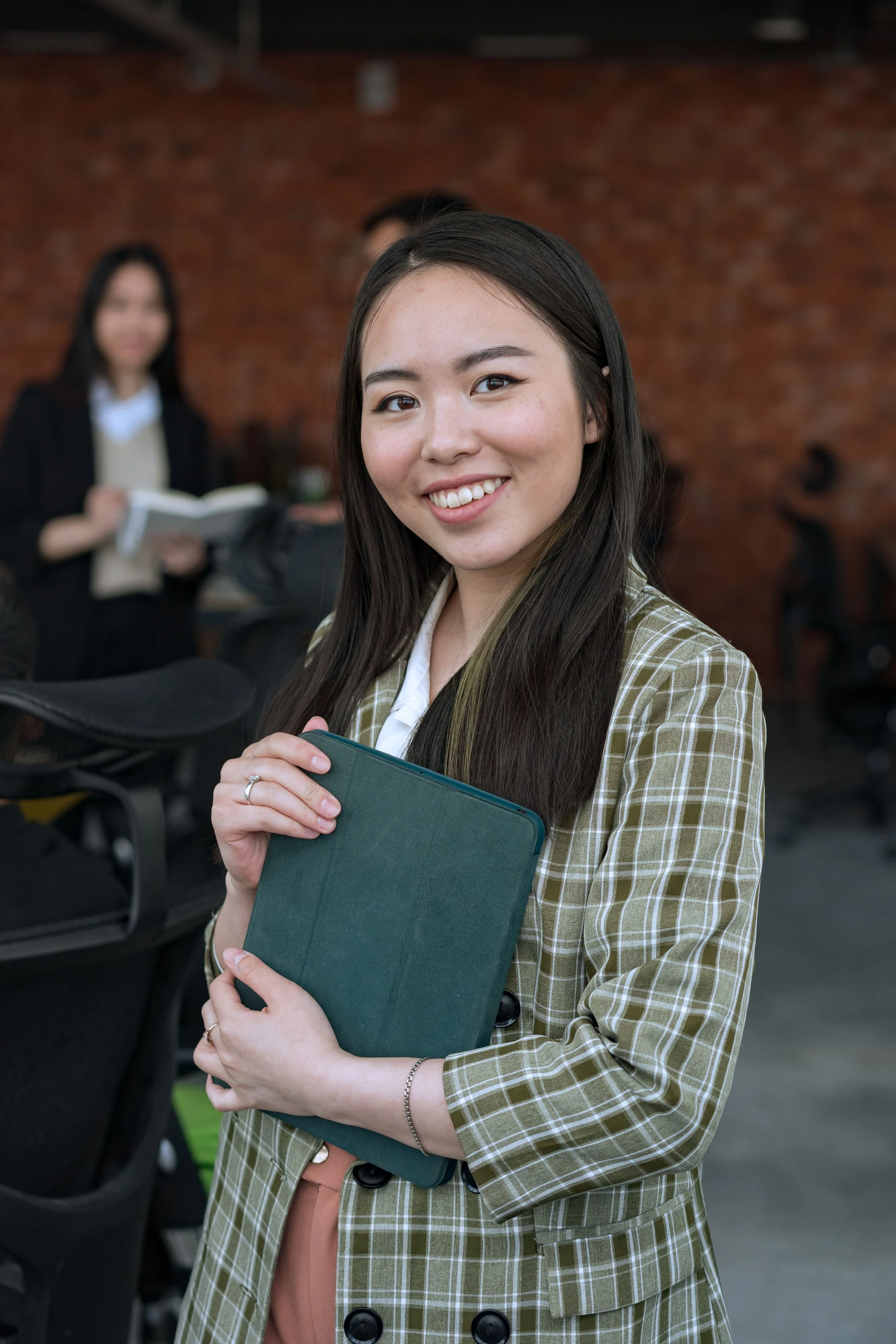 Young woman with long dark hair smiling, holding a dark green notebook, wearing a plaid jacket, in a modern indoor space with a brick wall background. Blurred woman in black blazer holding a book, and other people are visible in the background.