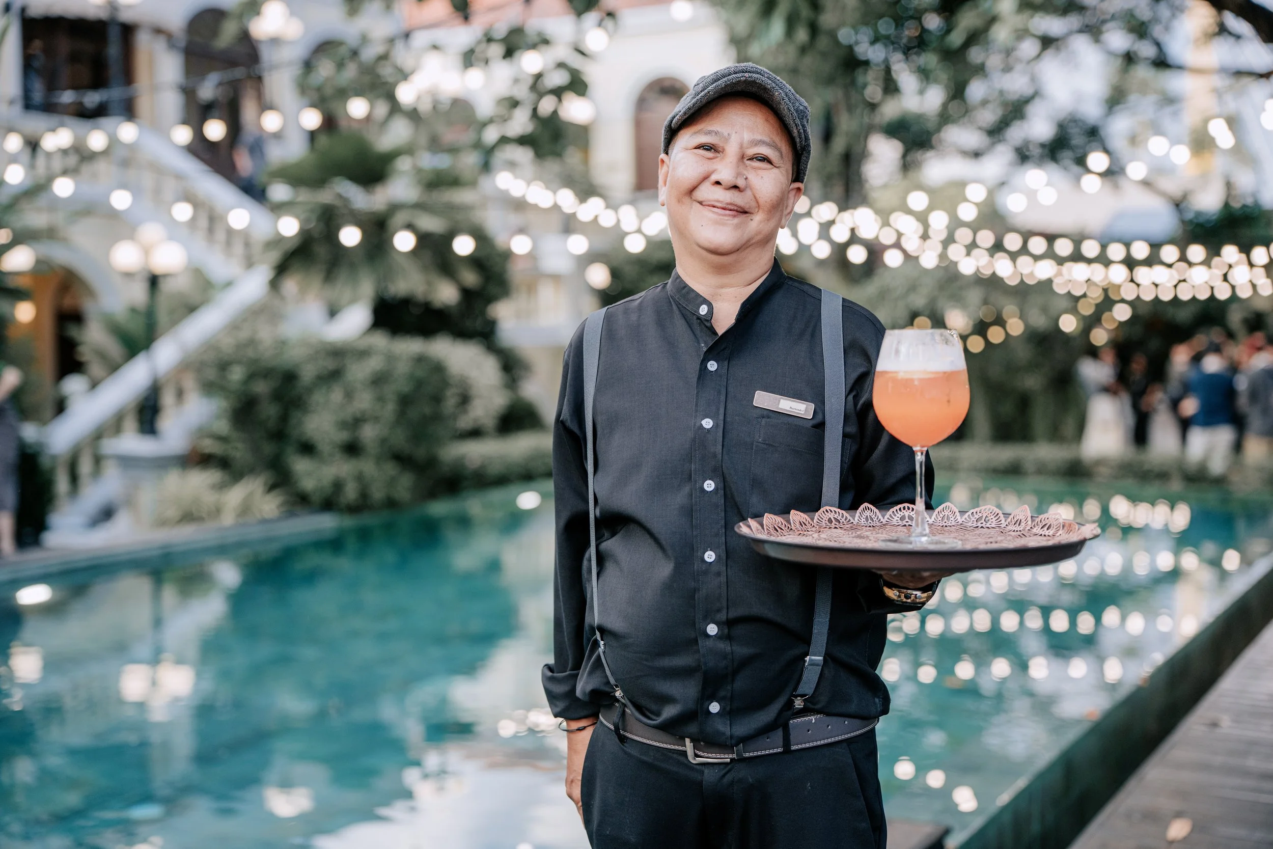 A smiling waitress wearing a black uniform and a gray cap holds a tray with a pink drink in a wine glass at an outdoor event. The background features a festive setting with string lights, trees, and a building, suggesting a lively social atmosphere.