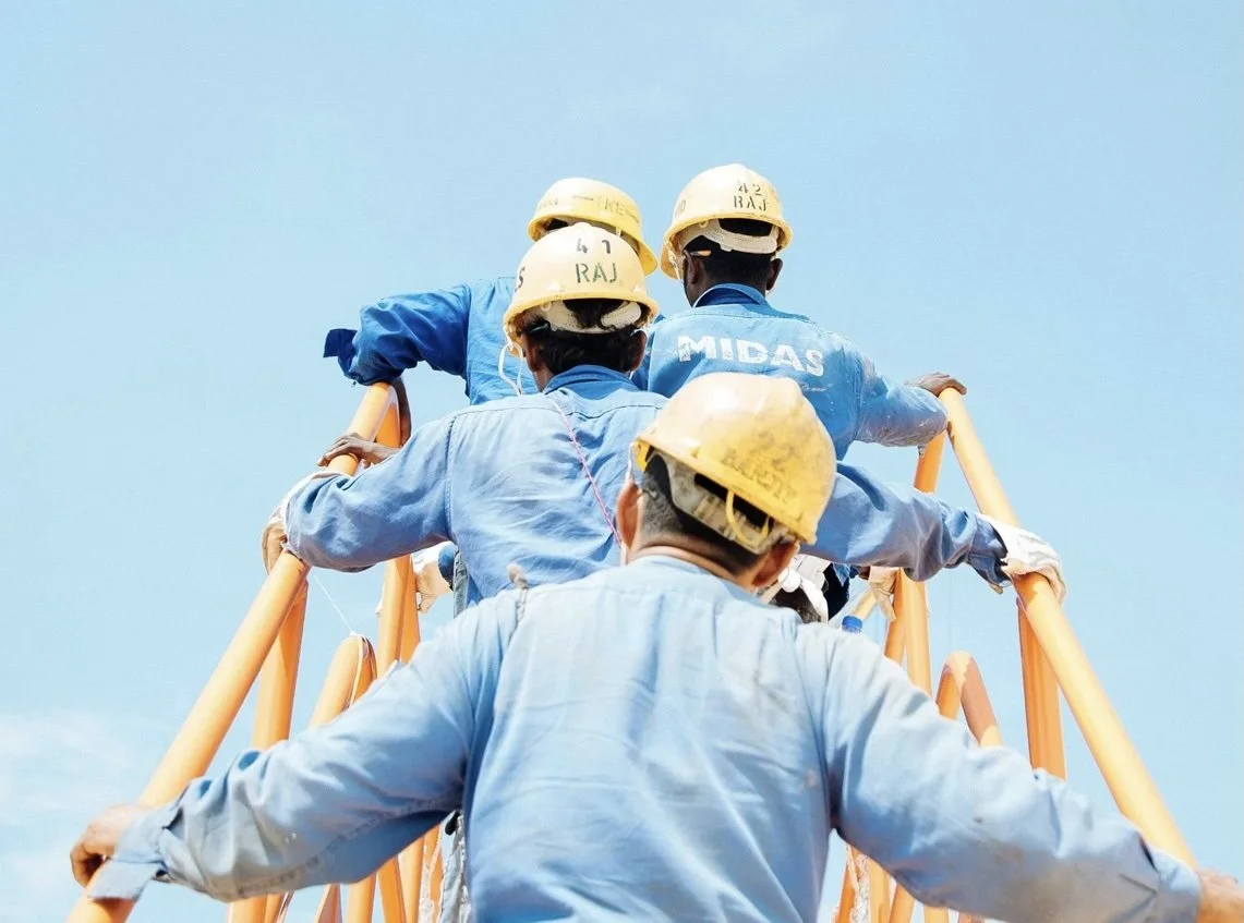Workers wearing yellow safety helmets climbing a tall orange ladder against a blue sky.
