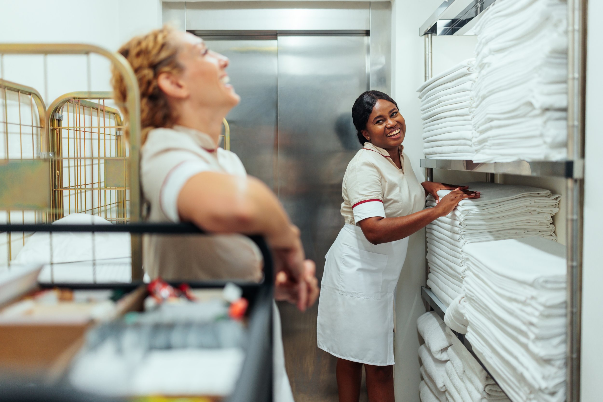 Two women in a commercial laundry room, one stacking folded white towels on a shelf and the other sitting on a laundry cart, smiling.