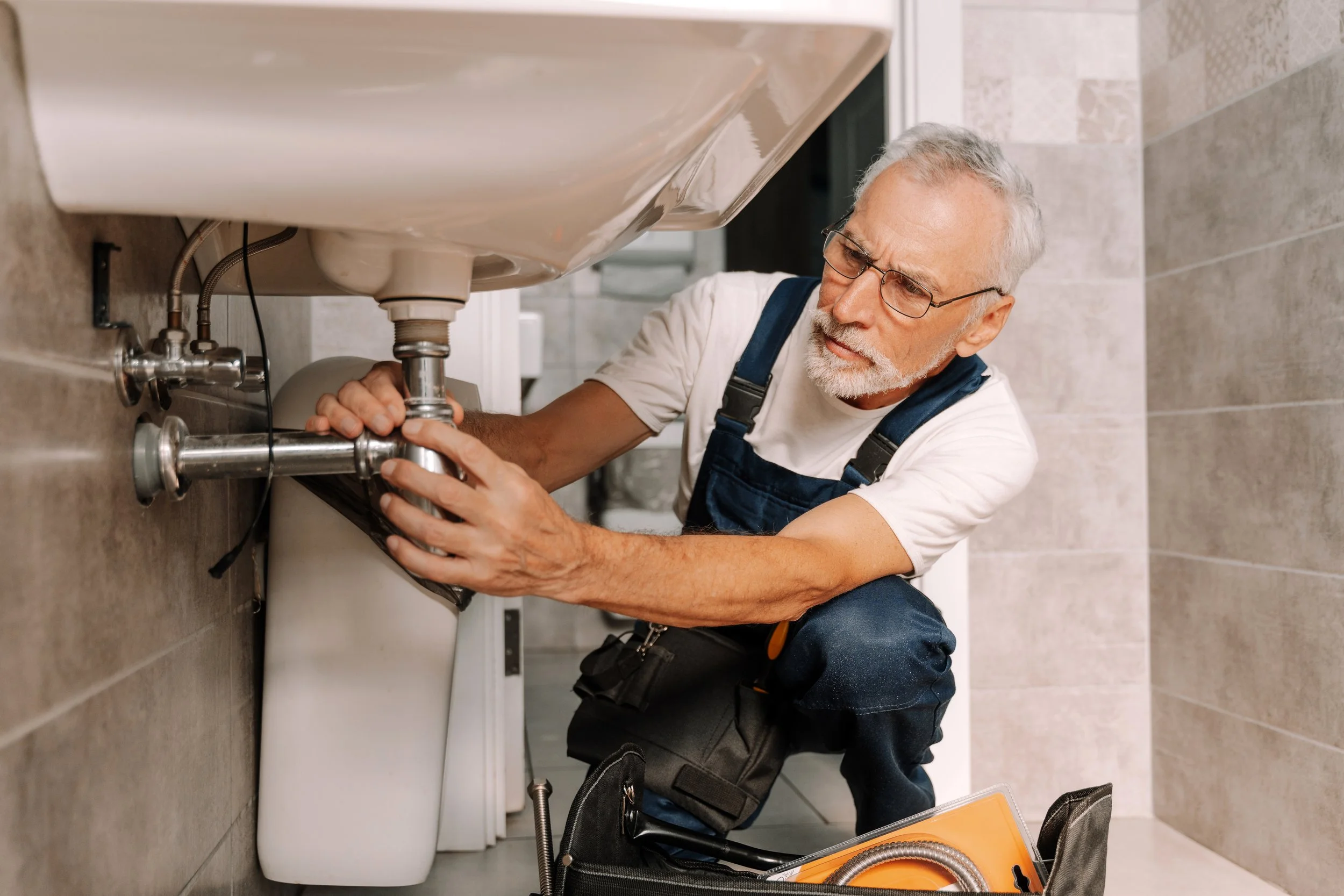 An elderly man with gray hair, glasses, and a beard fixing or inspecting the plumbing underneath a bathroom sink.