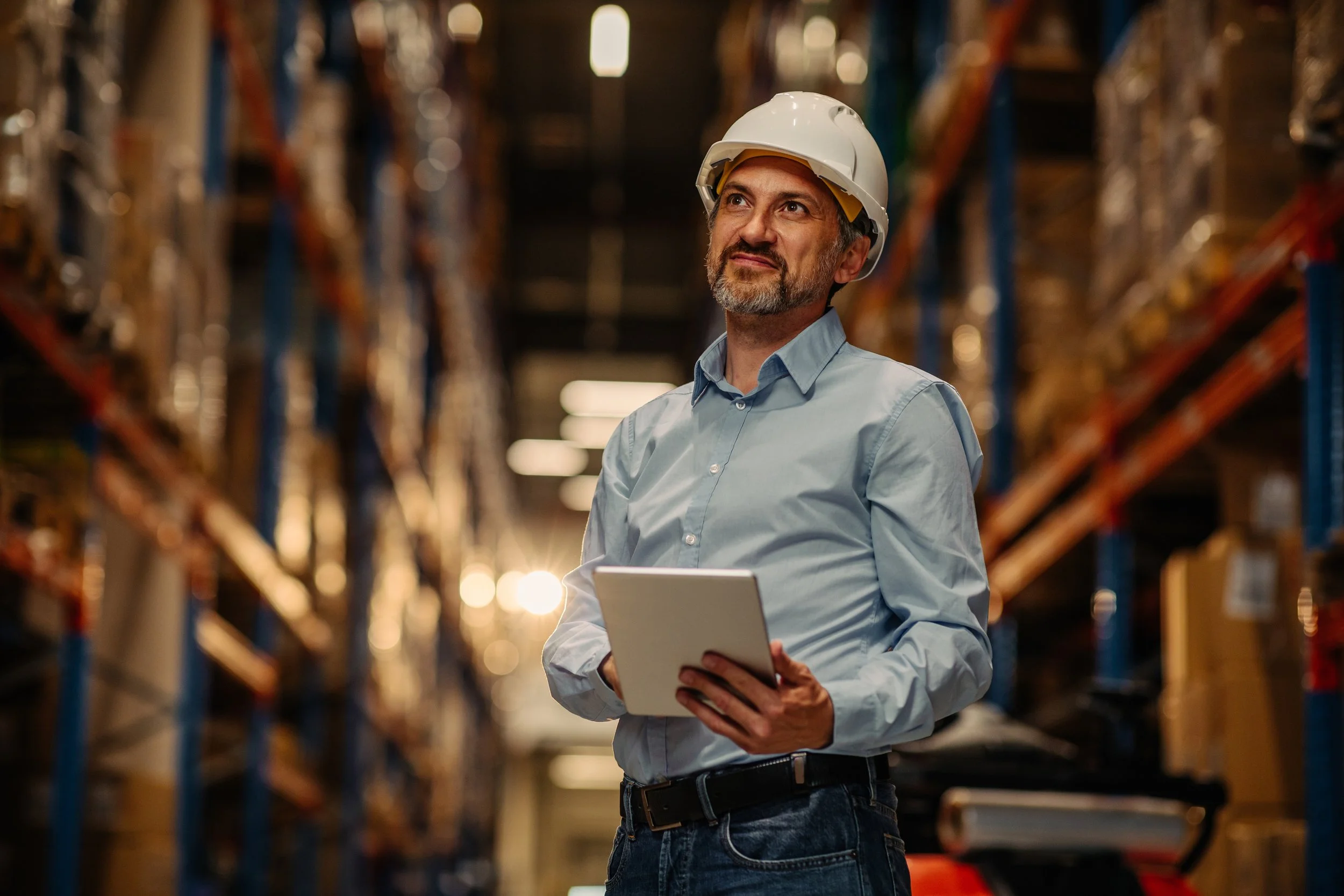 A man wearing a white safety helmet and a light blue button-up shirt, holding a tablet, standing in an aisle of a warehouse with shelves full of storage bins and equipment.