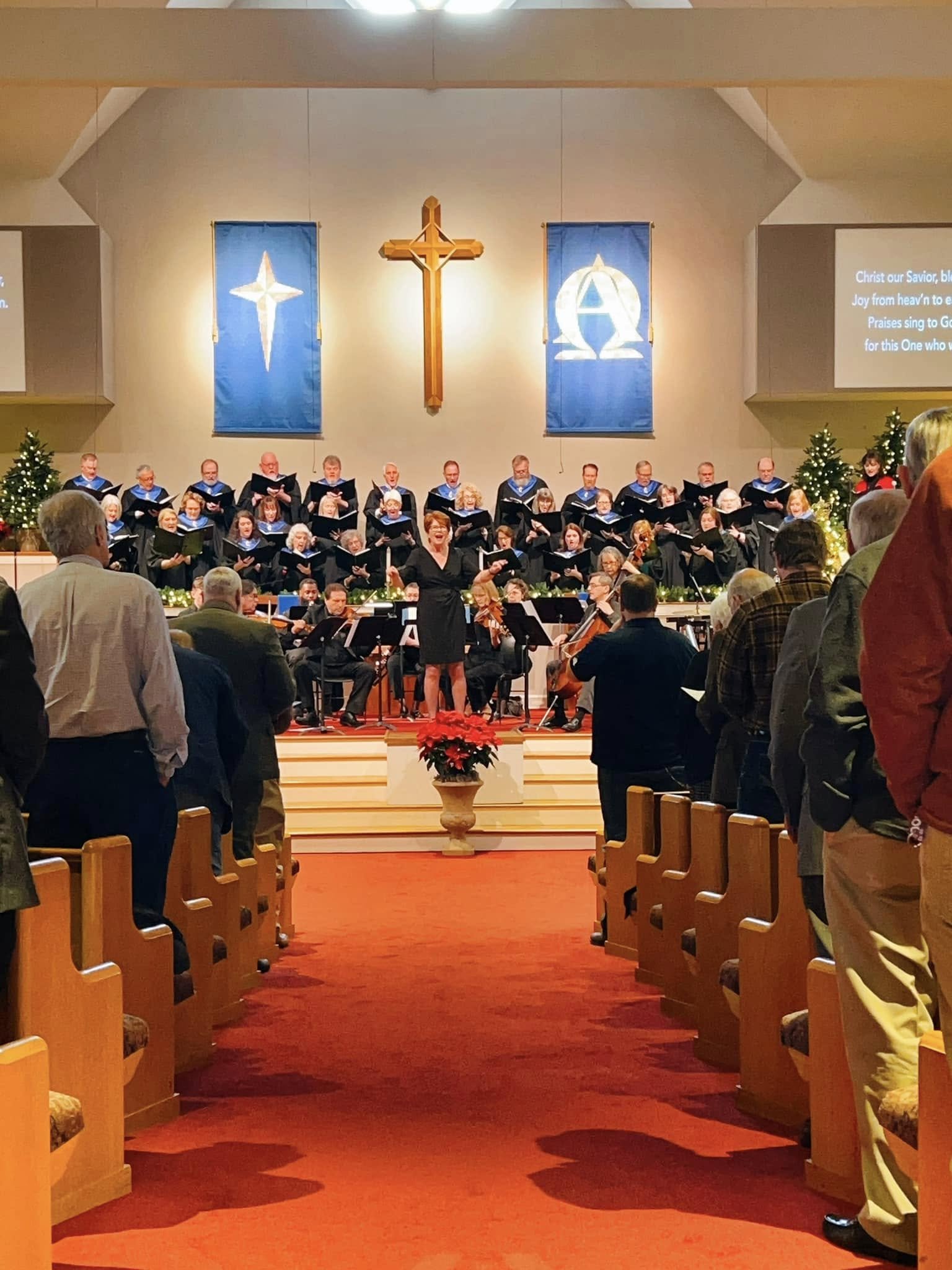 Choir performing on stage in a church during Christmas season. The choir members are dressed in black robes with blue collars, standing in front of blue banners with religious symbols. A woman in black conducts the choir, and the congregation watches. Christmas trees decorated with lights are on each side of the stage, and a poinsettia plant is in the foreground.