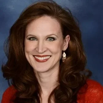 Portrait of a woman with long, wavy red hair, wearing a red top and earrings, smiling against a blue background.