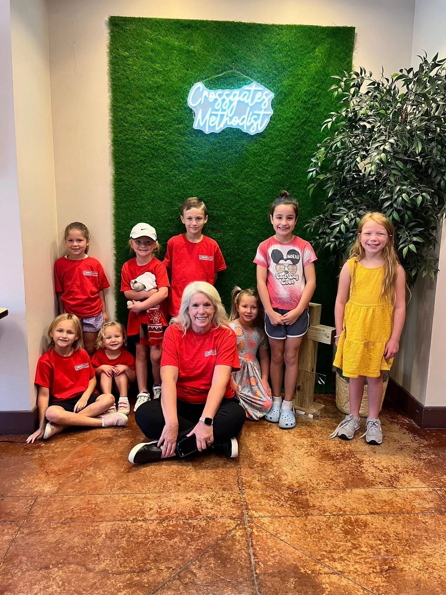 Group of children and two women posing in front of a green wall with a neon sign that says "Crossgates Methodist". The children are wearing casual and colorful clothes, some with red shirts. The women are sitting and kneeling on the floor, smiling at the camera.