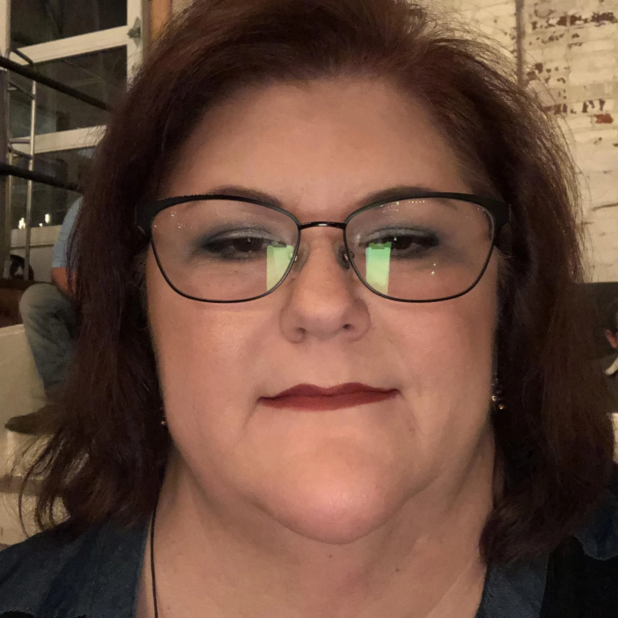Close-up of a person with curly reddish hair and glasses, wearing a black top, in an indoor setting.