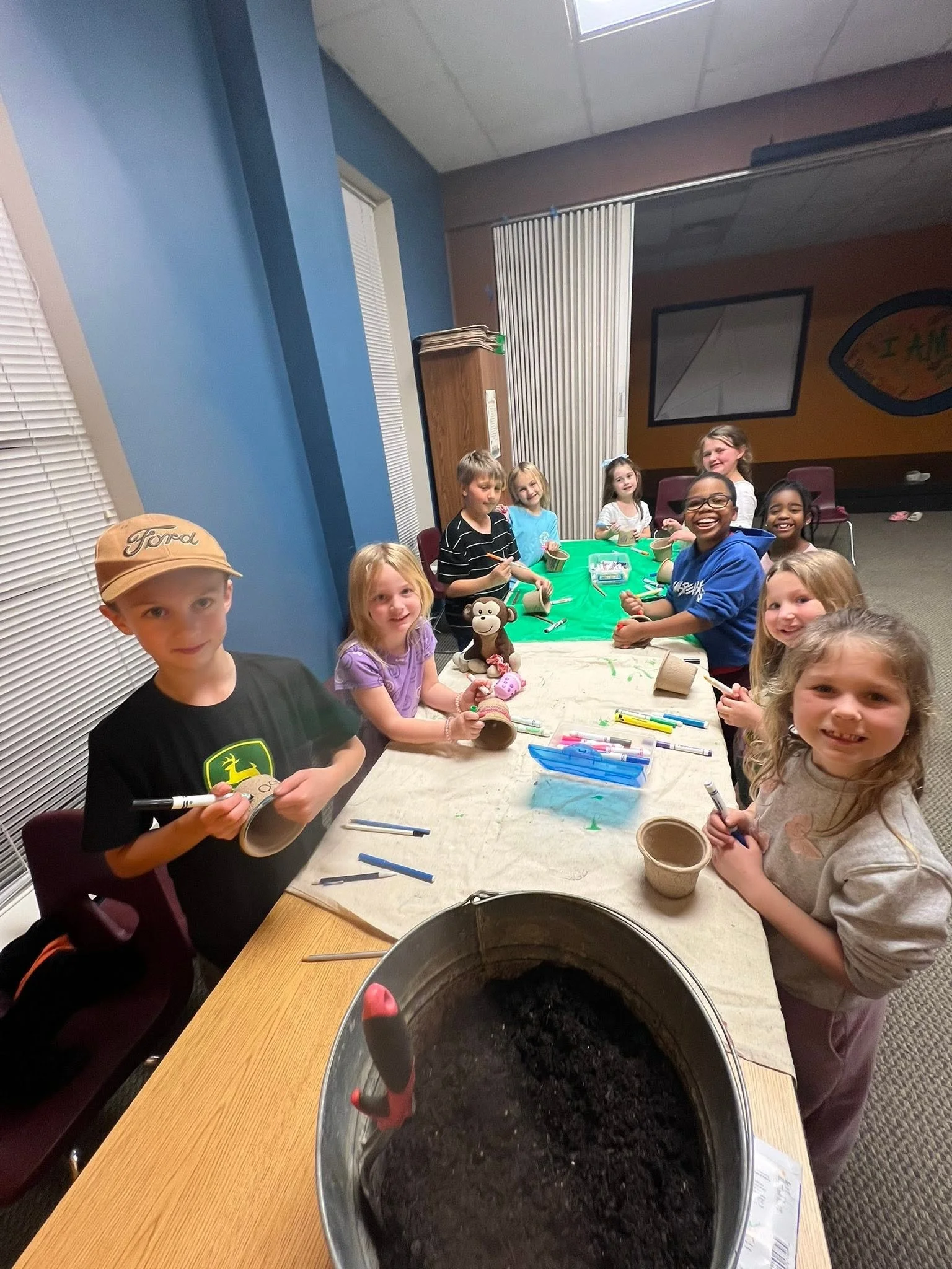 A group of children around a table, engaging in a craft activity, with some holding cardboard tubes and markers, and a large pot filled with soil in the foreground.