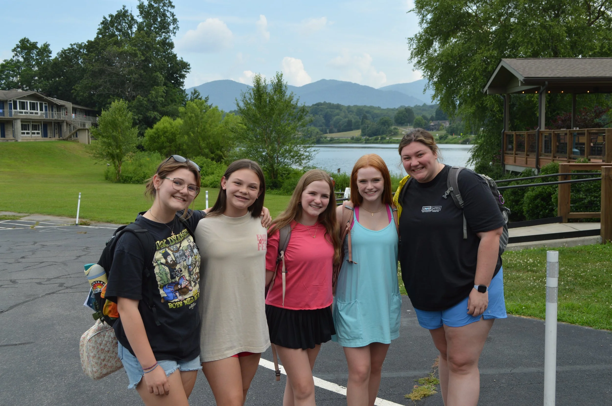 Group of five young women standing outdoors near a lake with mountains in the background, smiling at the camera, dressed casually, with a mix of hair colors and styles.