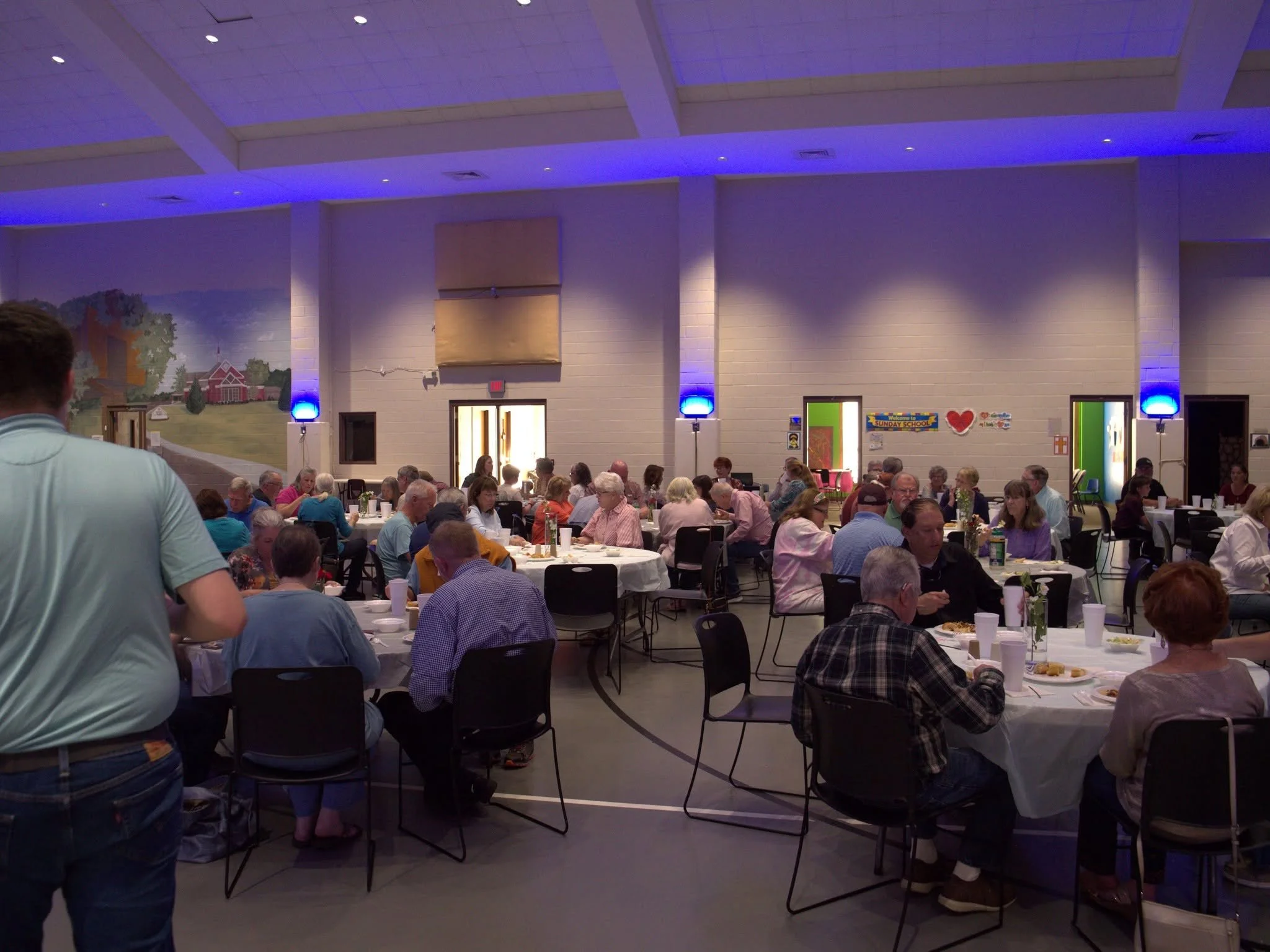 People sitting at tables in a large, well-lit room with white walls and a painted mural, attending a community event or gathering.