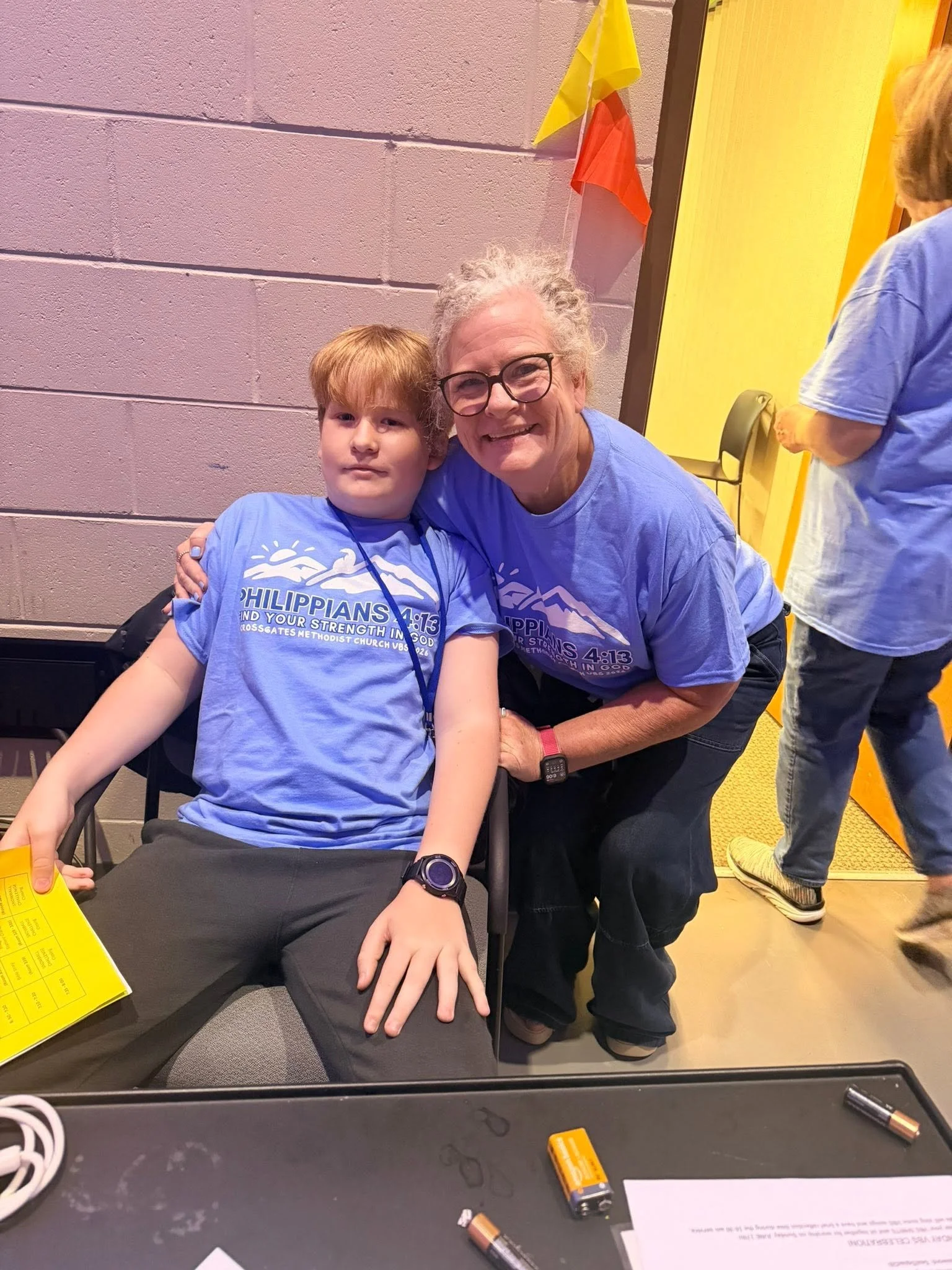 An older woman with glasses and a boy with red hair smiling at the camera. They are both wearing blue T-shirts with white text. The woman has her arm around the boy, and they are sitting at a table with some papers and scattered batteries. The background has a pink wall with colorful flags and another person partially visible wearing a light blue shirt.