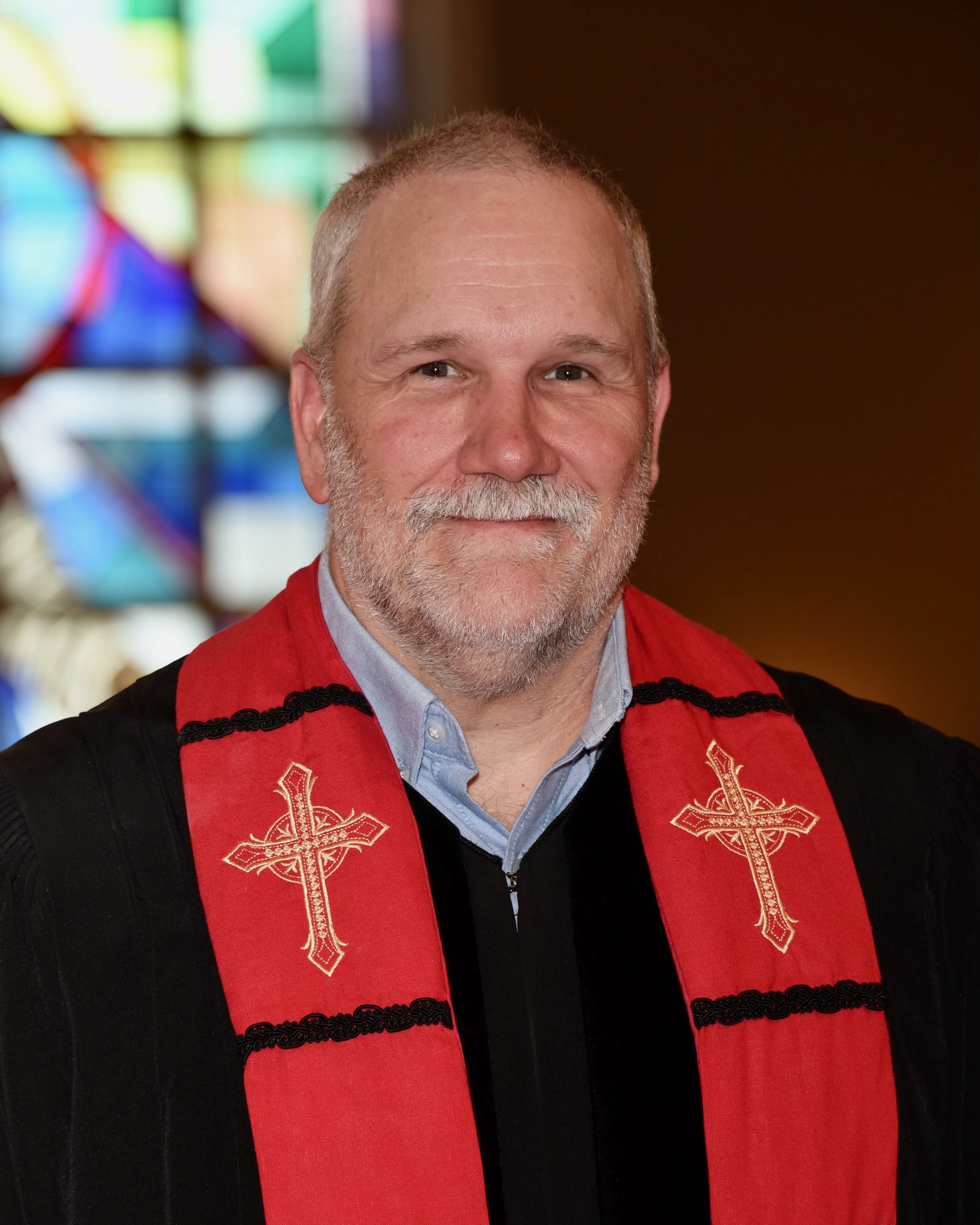 Close-up of a middle-aged man with gray hair and beard, wearing a black robe with red accents and a red stole adorned with gold embroidered crosses, standing inside a church with stained glass windows in the background.