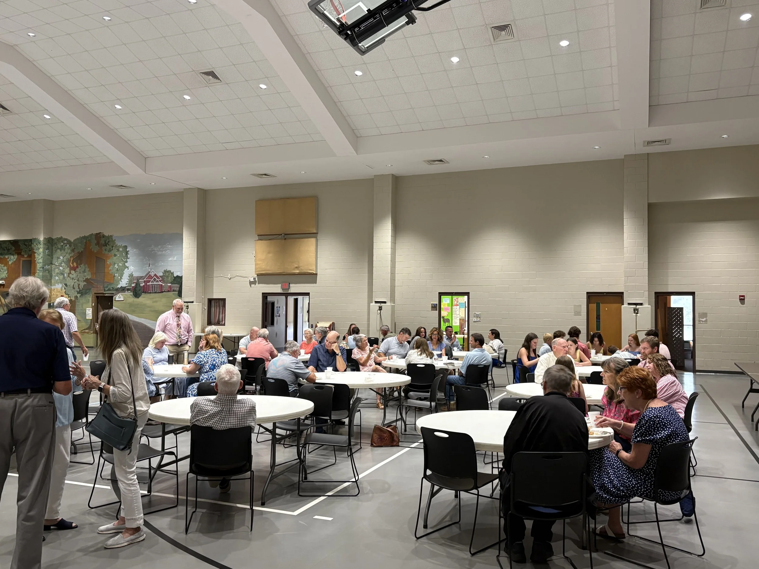 A large group of people seated at round tables in a community hall, engaged in conversations and eating. The hall has a high ceiling with bright lighting and a mural painted on one wall depicting a countryside scene.